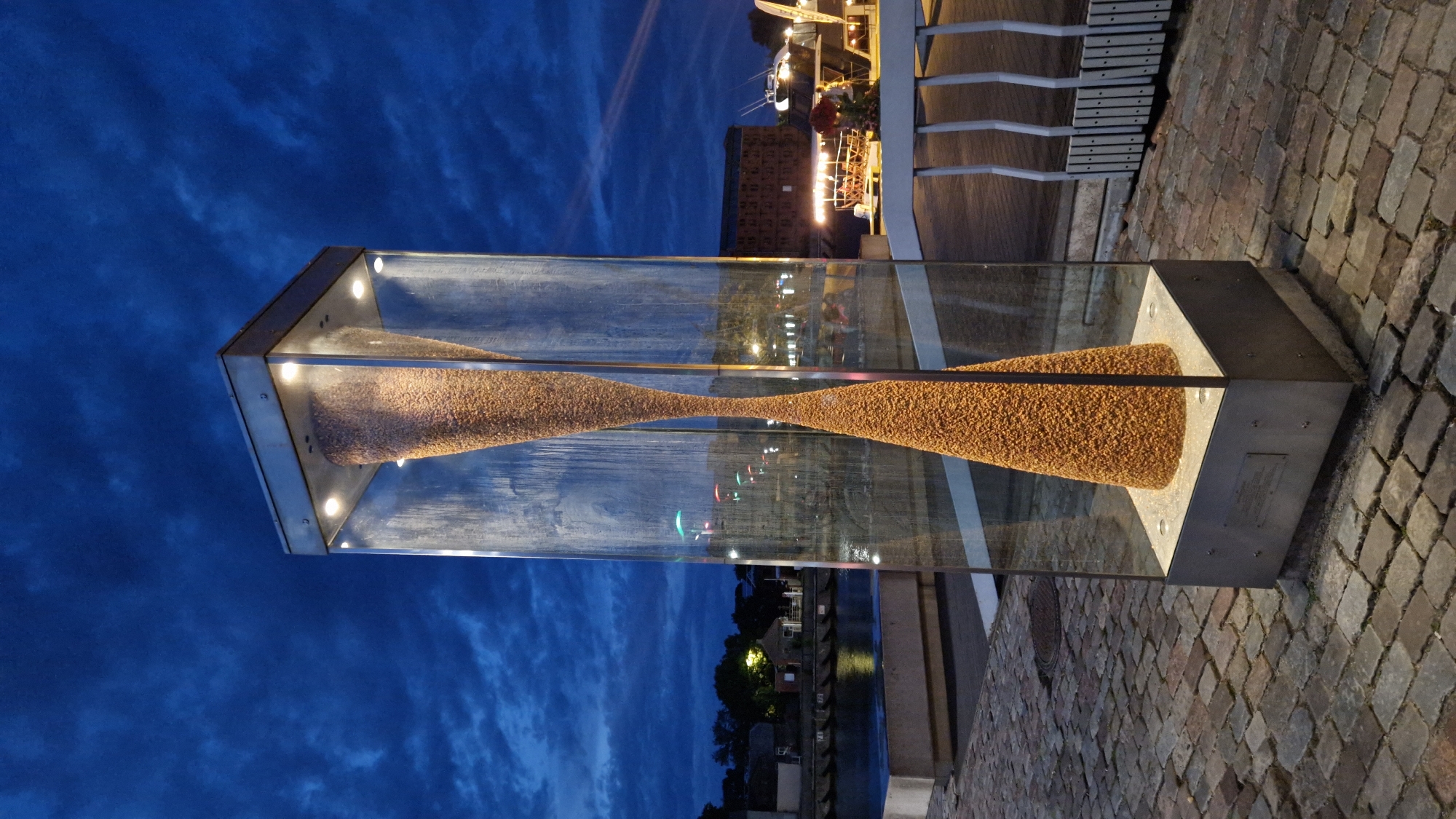 The image shows an outdoor sculpture resembling an hourglass filled with small, granular material, encased in a tall glass structure. The sculpture is illuminated from within, casting a warm glow on the material. It stands on a cobblestone surface in what appears to be a waterfront area. In the background, a body of water is visible, with buildings and boats illuminated by artificial lights. The sky is dark blue with clouds, suggesting it is either dusk or nighttime. The sculpture is located at the Liepaja Northern Breakwater in Liepaja, Latvia, specifically on the promenade. There are no people visible in the immediate vicinity of the sculpture, but distant figures and activity can be discerned in the background lights. The overall scene is one of urban waterfront art.