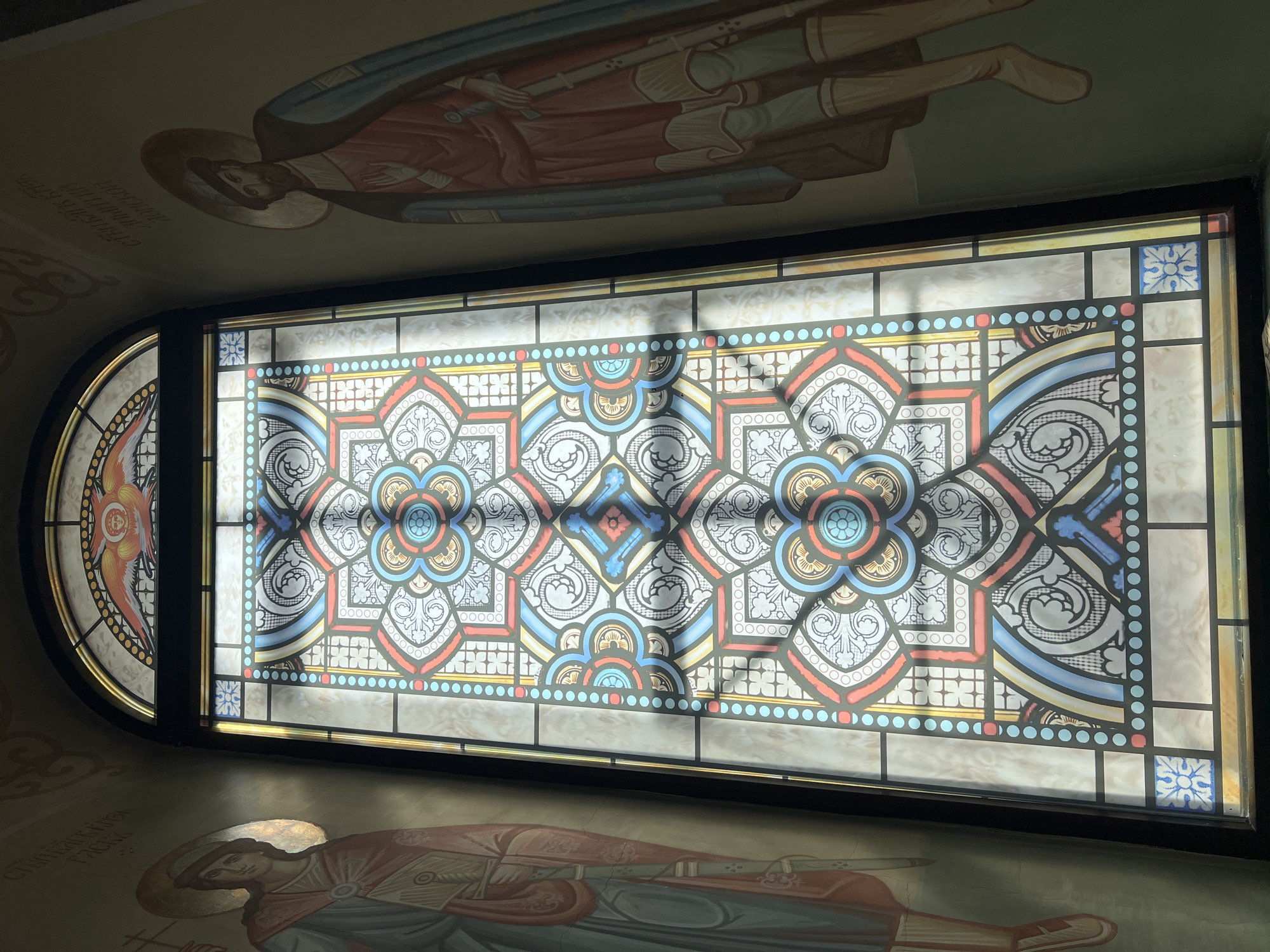 The image captures a stained-glass window within a church, likely in Kargapolye, Russia, given the style of iconography and architecture. The window is rectangular with a rounded section on the left. It is ornately decorated with geometric patterns and floral motifs, rendered in a variety of colors including blues, reds, yellows, and whites. The window is set in a dark frame, possibly made of wood or metal. Light filters through the window, creating shadows on the interior surfaces of the church. The top and bottom portions of the image reveal painted murals or frescoes on the ceiling and wall, depicting religious figures, with halo-like circles behind their heads. The overall impression is one of an Eastern Orthodox church interior.