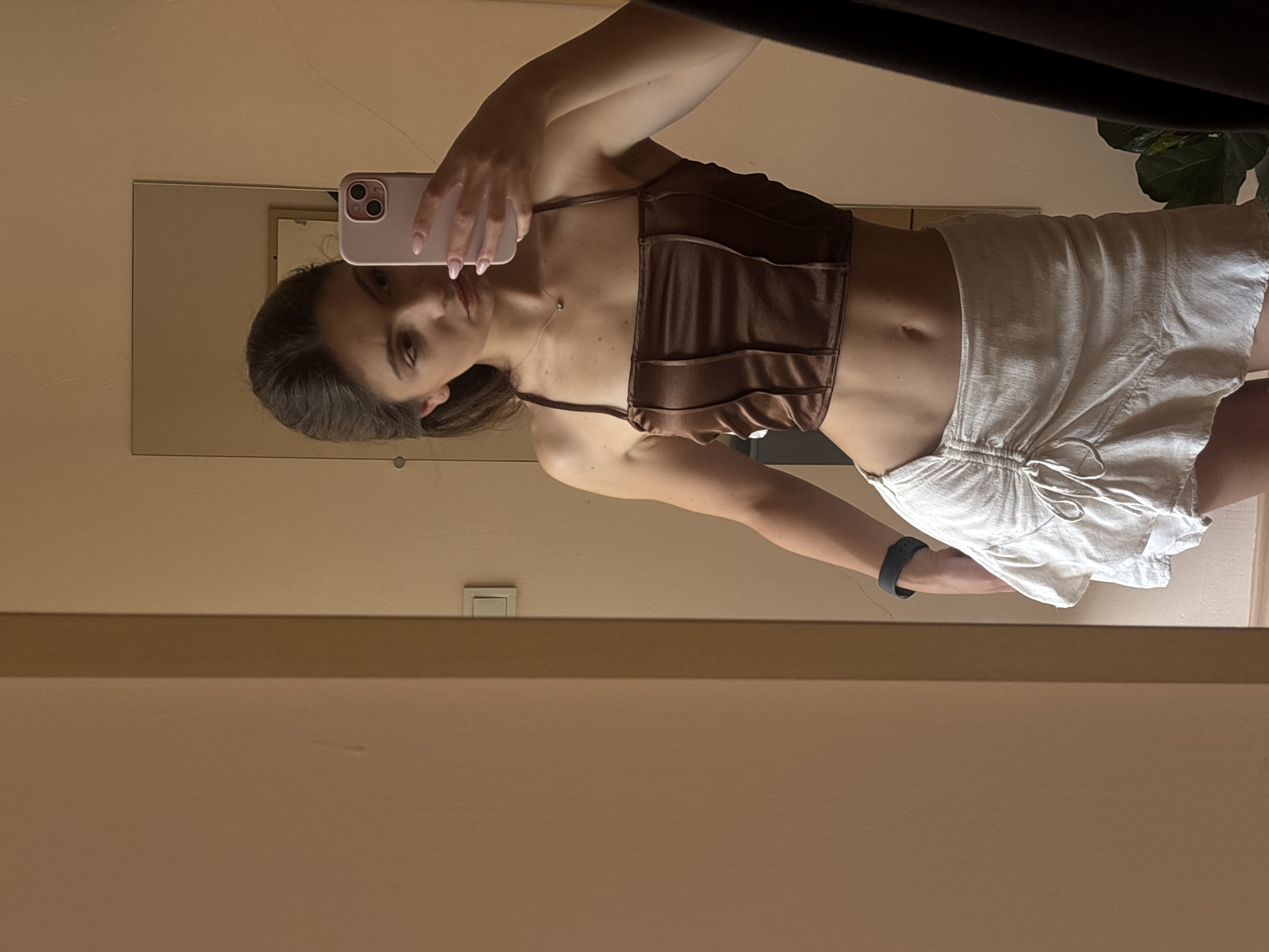 This is a vertical, selfie photograph taken in Varna, Bulgaria. The photo features a young woman with long brown hair, her face is partially visible as she holds a light pink smartphone to take the picture in a mirror. She is wearing a brown ruched halter top that exposes her midriff and a pair of loose, white shorts. Her left arm is visible, and she has a dark fitness tracker on her wrist. The background shows a neutral-colored wall and a mirror reflecting the woman and her surroundings. A corner of a dark piece of furniture and a green plant are visible at the top of the mirror's reflection. The lighting suggests it is daytime, possibly late afternoon or early evening, given the warm tones of the wall. The woman's expression is neutral, and the overall mood of the image is casual.