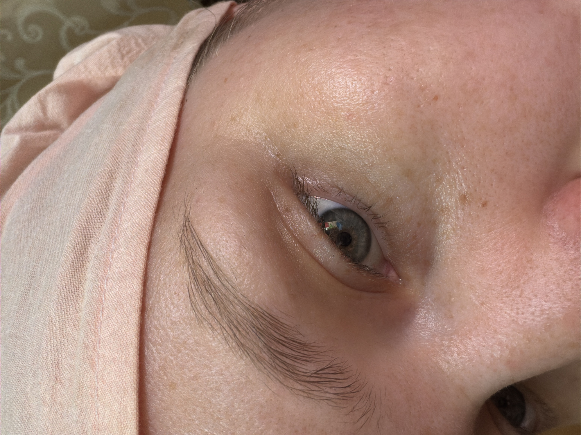 A close-up image captures a portion of a person's face, including the right eye, eyebrow, part of the nose, and forehead. The person is wearing a light pink headscarf. The eye is gray-green, and the eyebrow is relatively sparse. The skin is pale and has freckles. The background is out of focus, revealing a pattern suggesting a fabric surface. The location is not identifiable from the image.