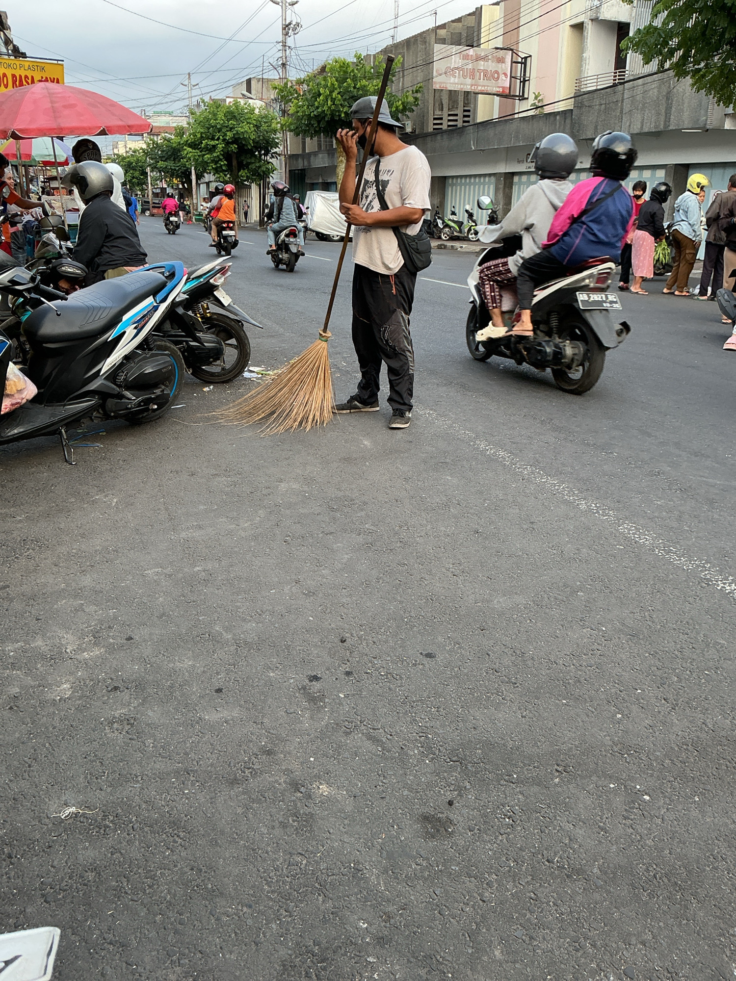 In Magelang, Indonesia, a street scene is depicted with a focus on transportation and daily life. In the foreground, a street sweeper, wearing a grey t-shirt, black pants, and a dark cap, holds a broom made of natural fibers. He appears to be sweeping debris near several parked motorcycles.
The background reveals a busy street with multiple motorcycles carrying passengers. One motorcycle in the mid-ground shows two individuals wearing helmets, one in a pink and blue jacket, and the other in grey. Another motorcycle further back has a rider in an orange shirt. Pedestrians are also visible on the sidewalk, some walking and others gathered near a food stall with a red umbrella. Buildings line the street, with signs indicating businesses. The sky is overcast, suggesting a cloudy day. The overall impression is of an active urban environment with a focus on pedestrian and vehicular movement, and the presence of street cleaning services.
