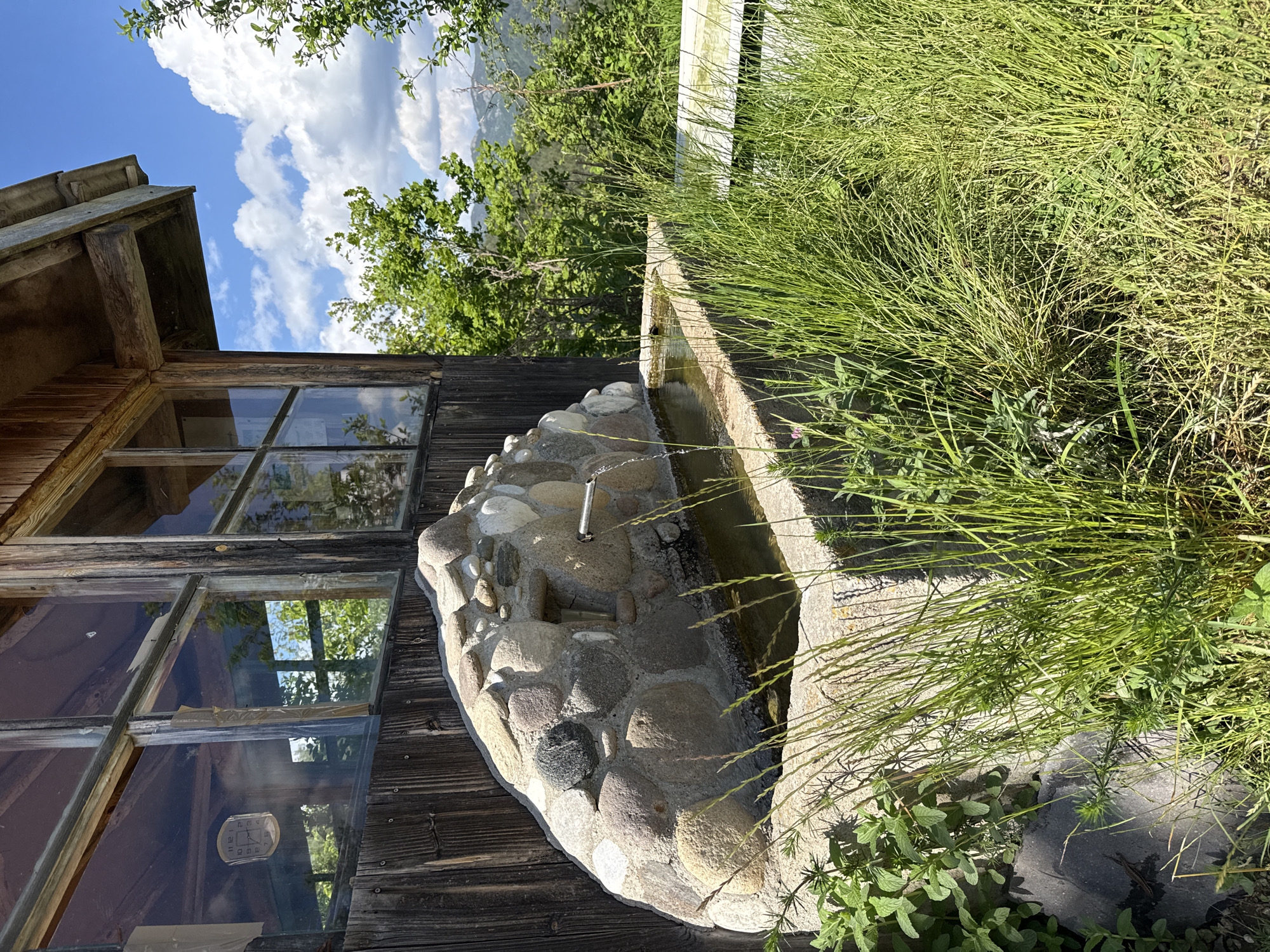 The image shows an outdoor scene in Ogled, Bulgaria featuring a rustic stone and concrete water trough with a metal spout, partially obscured by tall green grass. The trough is attached to the side of a weathered wooden structure with multi-paned windows. The windows reflect the sky and surrounding foliage. Behind the trough and structure, there is greenery and glimpses of mountains. The sky is blue with puffy white clouds. A white wooden fence is partially visible behind the grass. A clock is visible inside the window of the wooden structure.