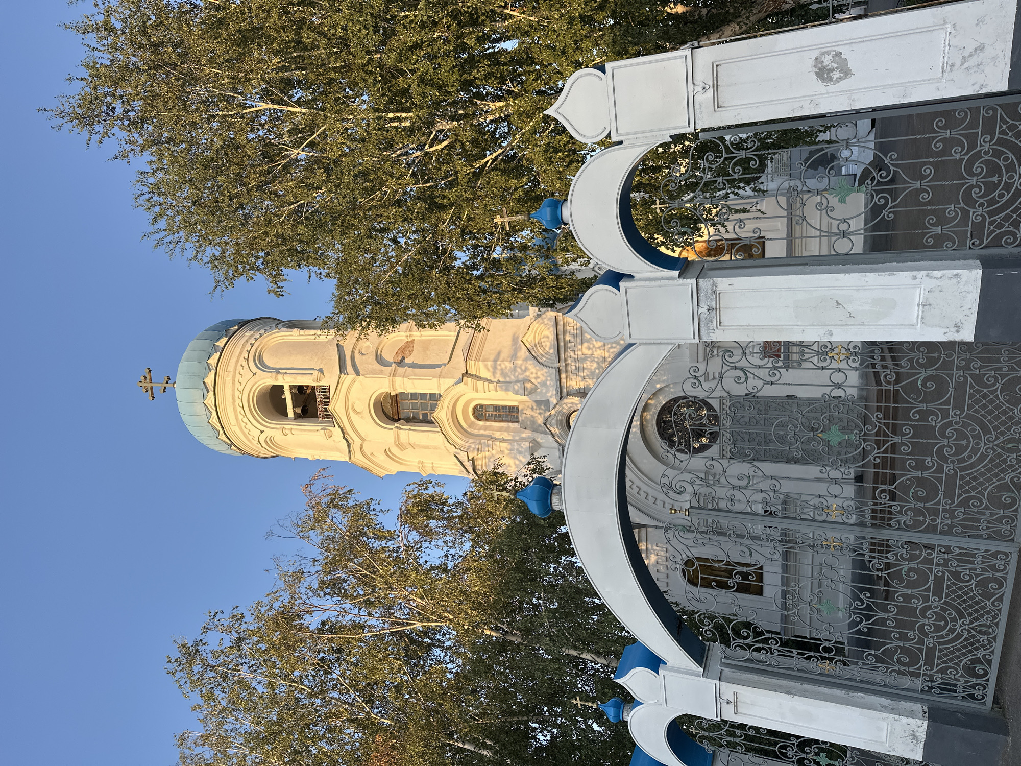 The image shows the bell tower and entrance gate of the Assumption Cathedral in Biysk, Russia. The bell tower is tall and white, with a blue dome and a cross on top. It has several arched windows and decorative elements. The entrance gate is made of white stone and grey metal with ornate patterns. It is flanked by white pillars with blue decorative elements and crosses on top. Behind the gate, the church building is visible, with more blue domes and white walls. Tall trees with green leaves surround the cathedral. The sky is clear and blue. The lighting suggests it is late afternoon or early evening, with the sun casting a warm glow on the buildings. There are no people visible in the image.