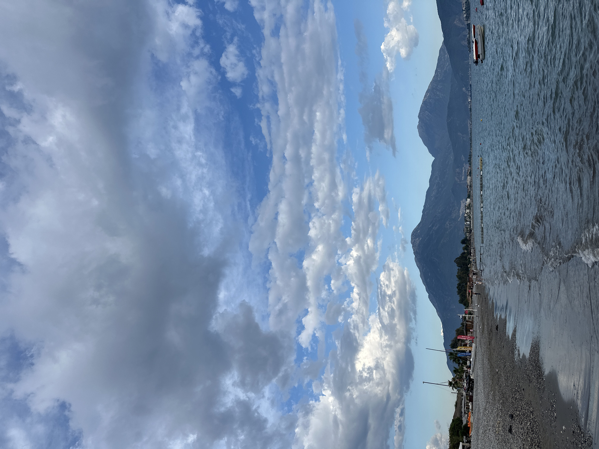 The image captures a view of a beach in Fethiye, Turkey, with a rocky shoreline. A mountain range forms the backdrop, meeting the sea. The sky is filled with a mix of white and grey clouds. A few boats are visible in the water, and beach umbrellas and flags dot the shoreline. The scene suggests a tourist destination, with people possibly enjoying the beach.