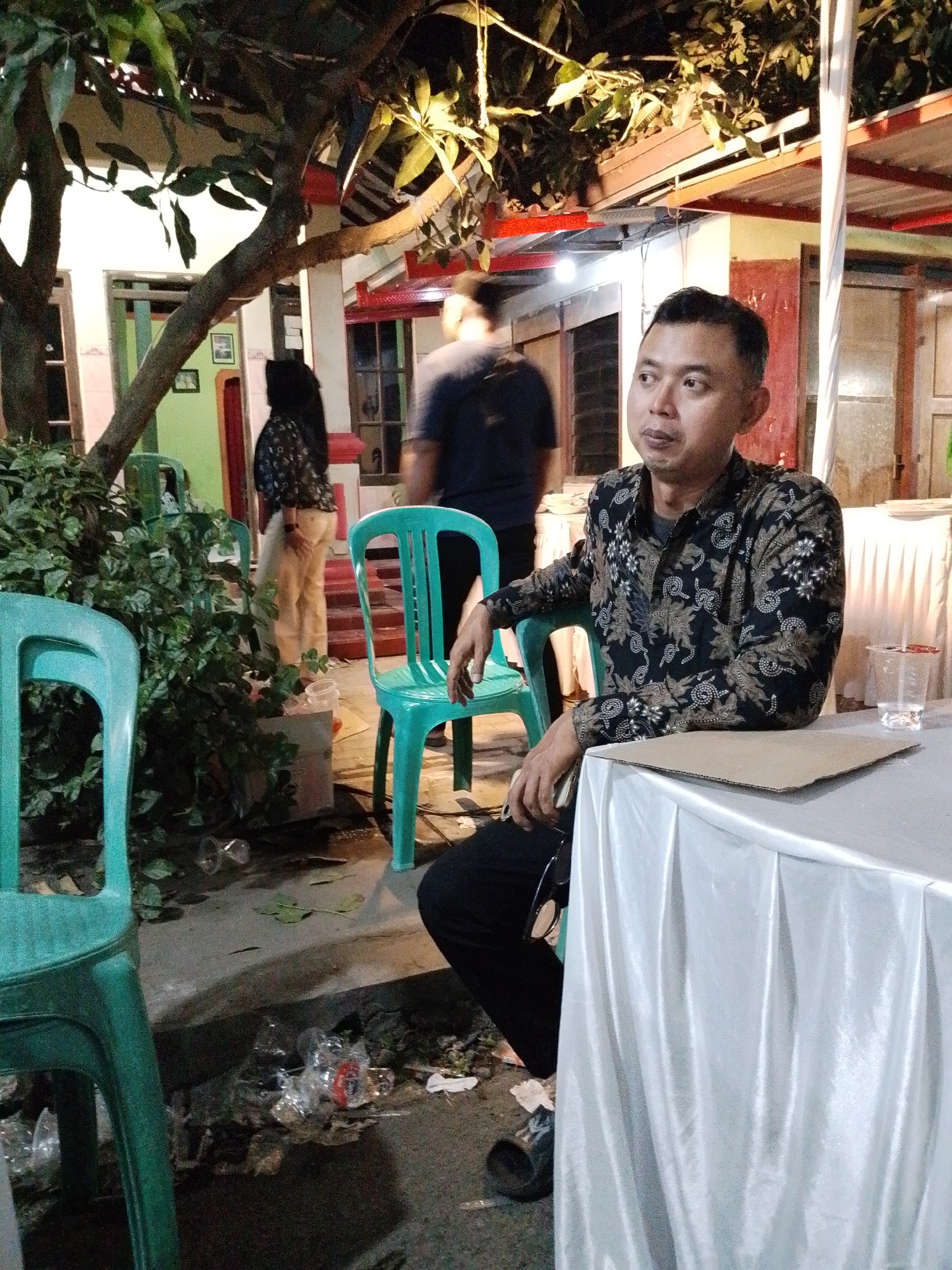 A medium shot captures a man in a patterned shirt sitting at a table draped with a white tablecloth, outdoors in Trangkil, Indonesia. The man is looking to his right, with his left arm resting on a green plastic chair. In the background, a woman in a headscarf and patterned shirt stands near a building entrance. Other green plastic chairs are visible, along with scattered debris on the ground, including plastic bottles and wrappers. Trees with foliage hang over the scene, and artificial lighting illuminates the area. The overall environment suggests an outdoor gathering or event.