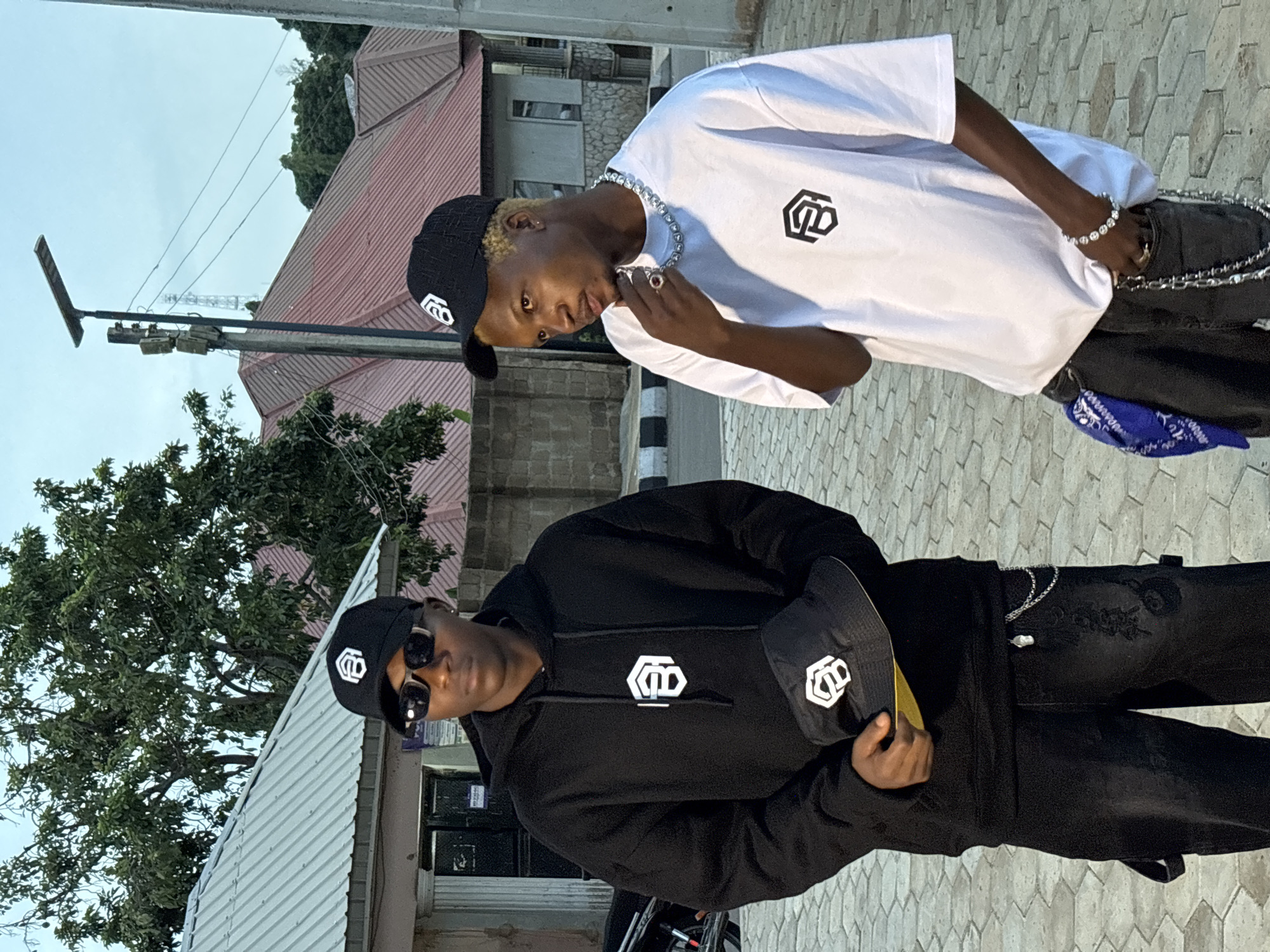 Two young men are standing outdoors in what appears to be a residential or commercial area in Jos, Nigeria. The man on the left is wearing a black hoodie with a white logo on the chest, black pants, a black baseball cap, and sunglasses. He is holding a black bucket hat with a white logo. The man on the right is wearing a white t-shirt with a black logo on the chest, black pants, and a silver chain necklace. He has blonde-dyed hair. In the background, there are buildings with red roofs, trees, a street light, and power lines. The ground is paved with hexagonal stones.