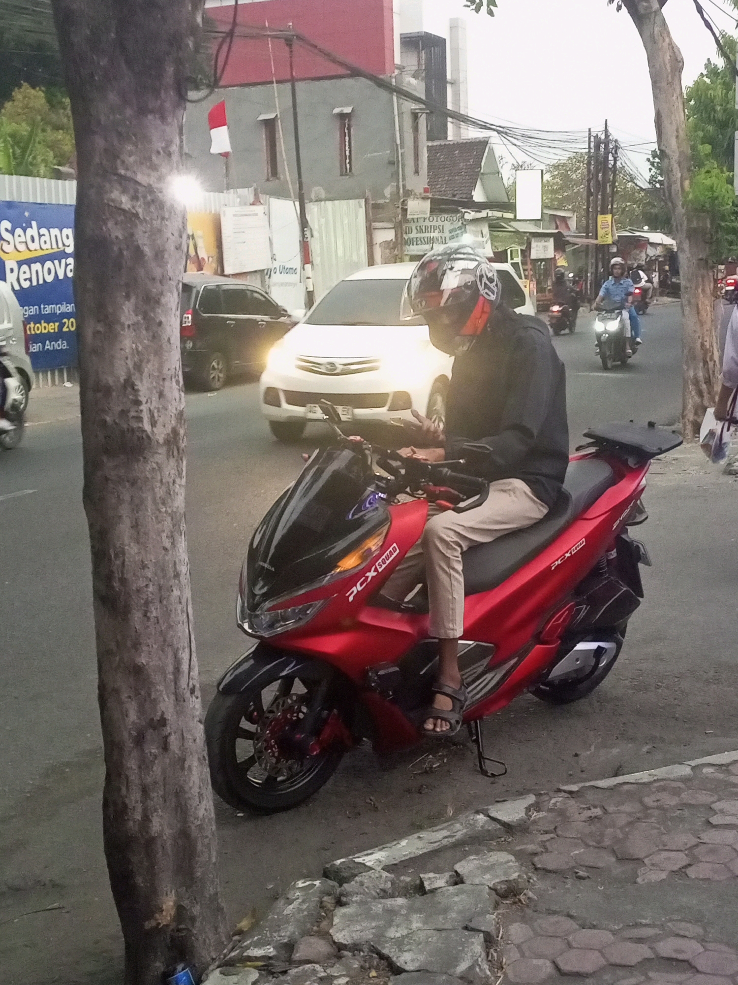 A person wearing a helmet and a black jacket is seated on a red scooter parked on the side of a street in Kediri City, Indonesia. The scooter has "PCX SQUAD" written on its side. The person is wearing light-colored pants and sandals. In the background, a white car and a dark-colored SUV are visible, along with other motorbikes. There are buildings and power lines lining the street. A tree trunk is prominently positioned in the foreground to the left of the frame. A blue banner with Indonesian text is also visible near the tree.
