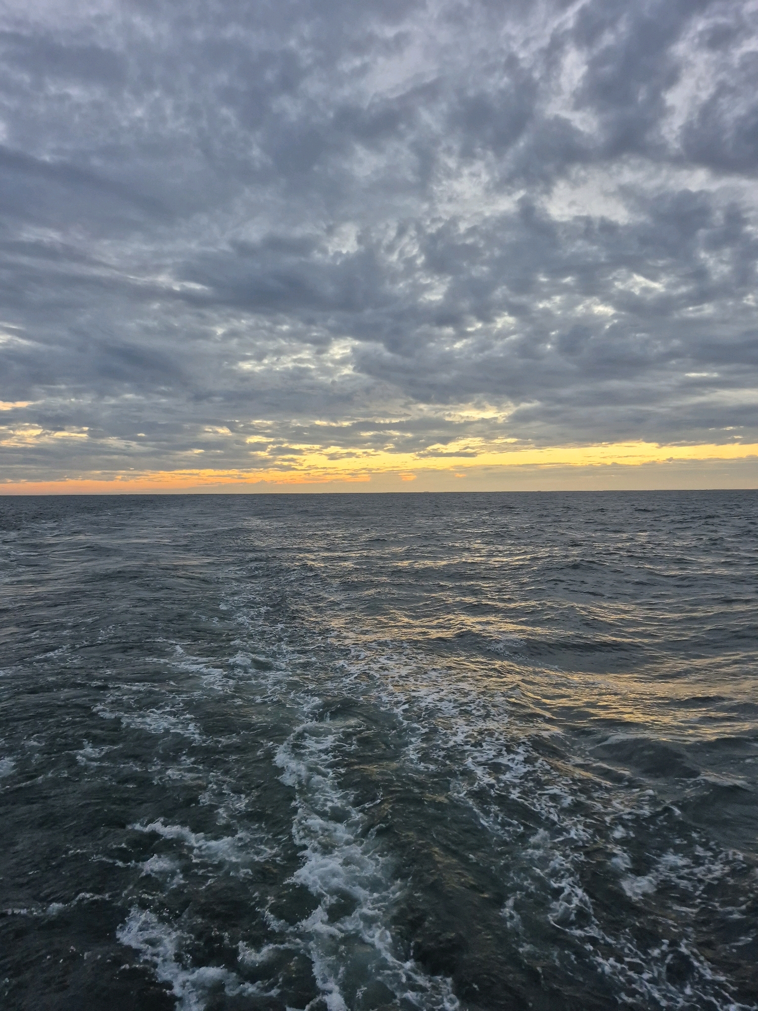 The image shows a wide view of the ocean at dusk. The sky is filled with heavy, gray clouds, with a band of orange and yellow light visible on the horizon, indicating the setting sun. The ocean surface is choppy, with white foam trailing behind the viewer, suggesting movement. The overall atmosphere is somber and tranquil. There are no people, buildings, or specific landmarks visible that would indicate a location in the city of Unknown, Germany. The scene is a natural landscape.