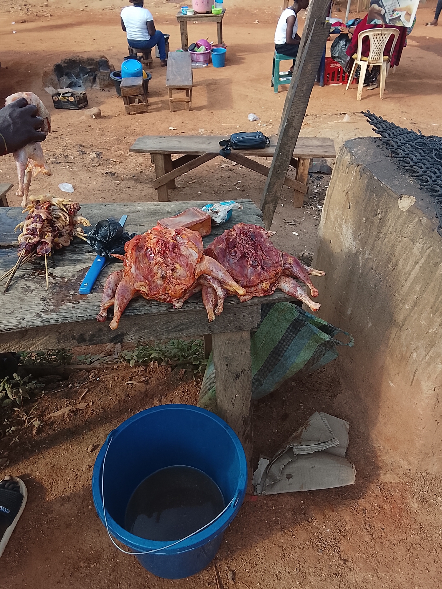 A high-angle medium shot captures an outdoor market scene with raw poultry prominently displayed on a weathered wooden table. Two whole chickens, freshly prepared and appearing to be cleaned, are positioned centrally on the table, their skin glistening and internal organs exposed. To the left, skewers of smaller pieces of meat, possibly chicken wings or gizzards, are arranged, suggesting a variety of offerings. A blue-handled knife and a black plastic bag lie on the table near the poultry. The table itself is rustic, made of rough-hewn planks, and sits outdoors on a dirt ground. In the background, out-of-focus figures are visible, including a person seated on a stool and another standing, possibly engaged in selling or preparing food. Various objects, such as buckets, chairs, and a grill-like structure, are scattered around the area, indicating a functional marketplace setting. The overall environment appears to be informal and possibly rural, with the ground covered in reddish-brown dirt and small pebbles.