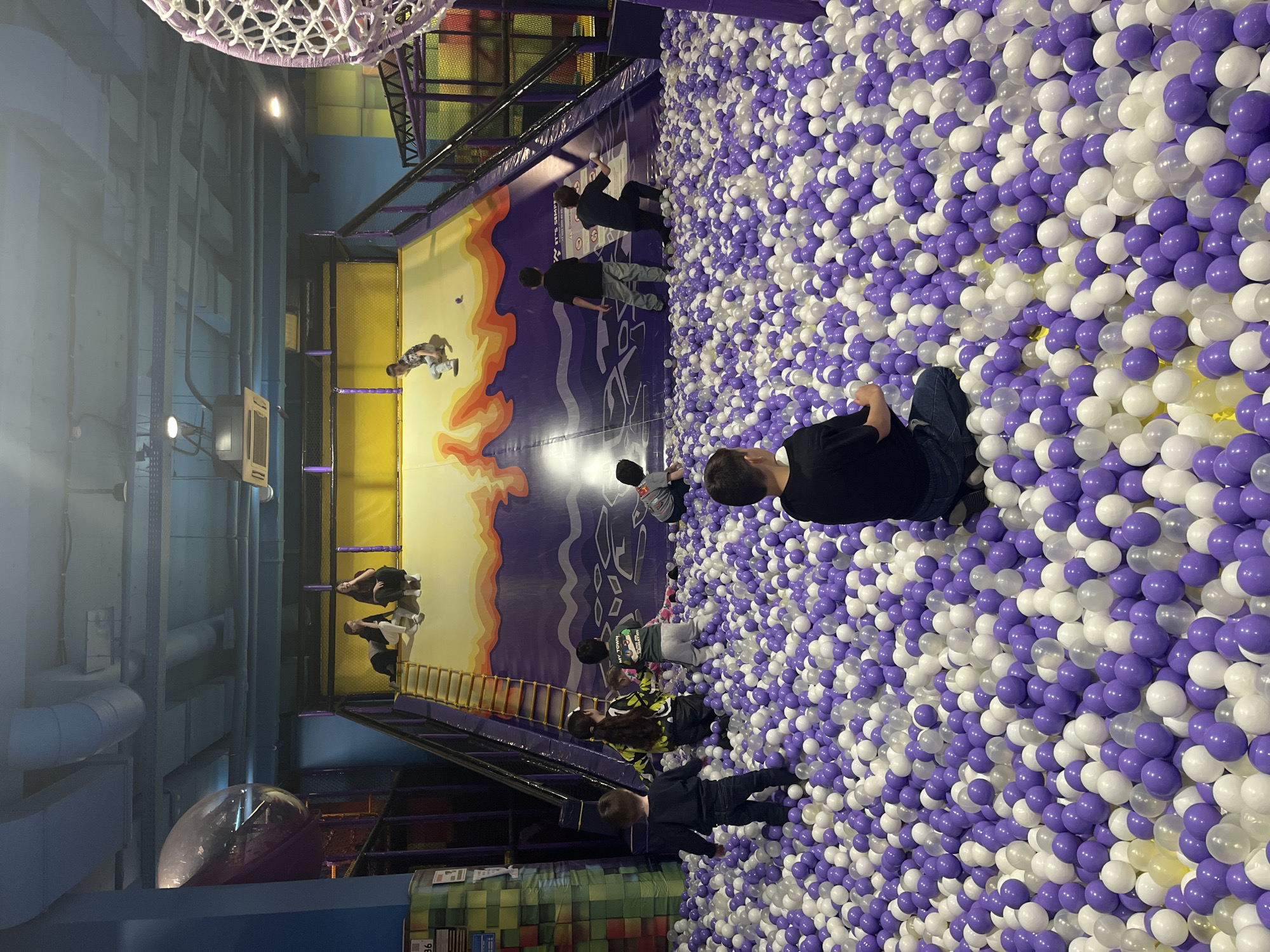 The image shows an indoor playground ball pit in Sochi, Russia. The pit is filled with white and purple plastic balls. Several people, including children and adults, are in the ball pit. Some people are sitting, while others are standing. The walls of the playground are decorated with images of landscapes and cityscapes.