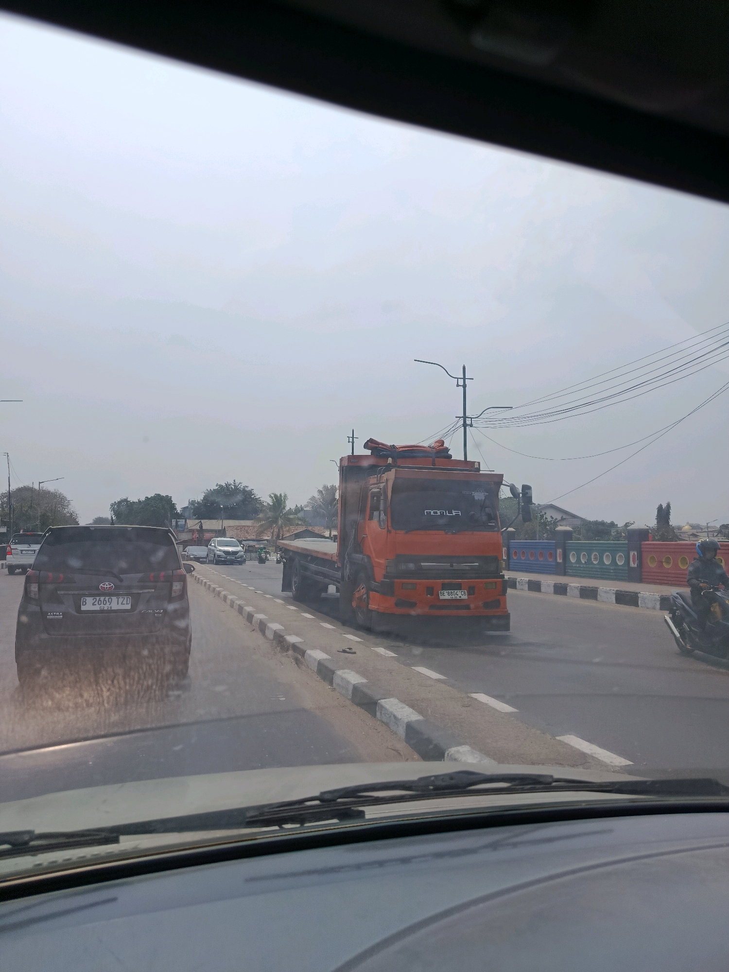 The image captures a view from inside a vehicle looking out onto a street in East Jakarta, Indonesia. The sky is overcast, creating a hazy, diffused light. A black car, with the license plate B 2669 TZD, is in the foreground, partially obscuring the view of the road ahead. To the right of the black car, a bright orange truck is driving in the same direction. The truck has a visible license plate that appears to read "BE 8804 CB". It is carrying some cargo on its flatbed. Further down the road, a person on a motorcycle, wearing a helmet, is also moving in the same direction. The street is lined with a sidewalk that has a decorative barrier painted with various colors. Trees and buildings are visible in the background, suggesting an urban environment. Traffic lights and power lines are also present.