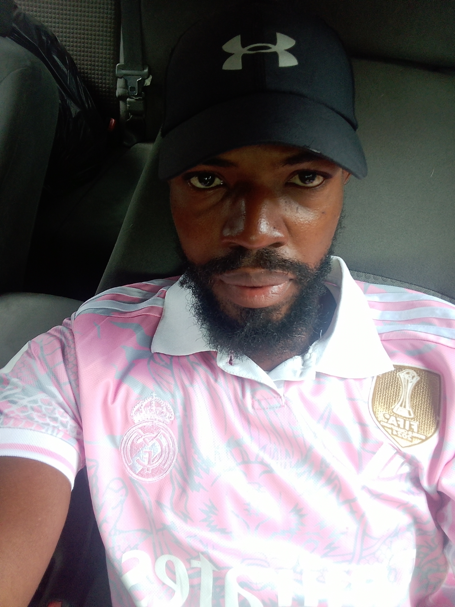 A man with a beard, wearing a black baseball cap with a white logo, and a pink and white patterned t-shirt, is sitting in a vehicle in Iju, Nigeria. The man is looking at the camera. The background shows the interior of the vehicle, including a seatbelt and a dark-colored seat.
