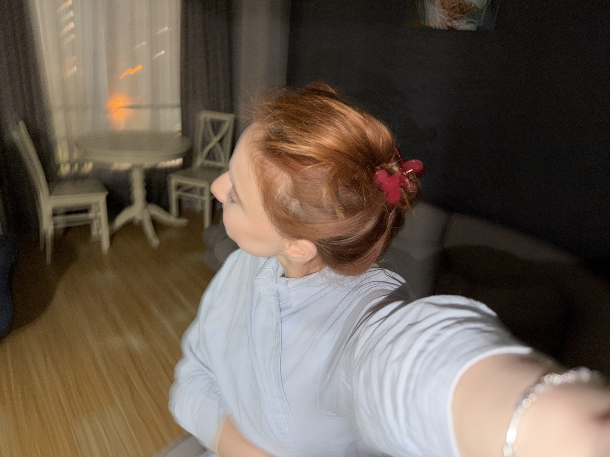 The image appears to be a selfie taken indoors. A woman with reddish-brown hair pulled back with a red hair clip is the main subject. She's wearing a light blue, button-down shirt. A silver bracelet is visible on her wrist.
In the background, there is a dining area with a round table and two white chairs near a window with sheer, light-colored curtains. Outside the window, blurred lights suggest a city environment. To the right of the woman is a dark gray sofa. The lighting in the room is a mix of natural light from the window and artificial light.
The image provides no concrete information to pinpoint the location to the city of Tbilisi, Georgia.