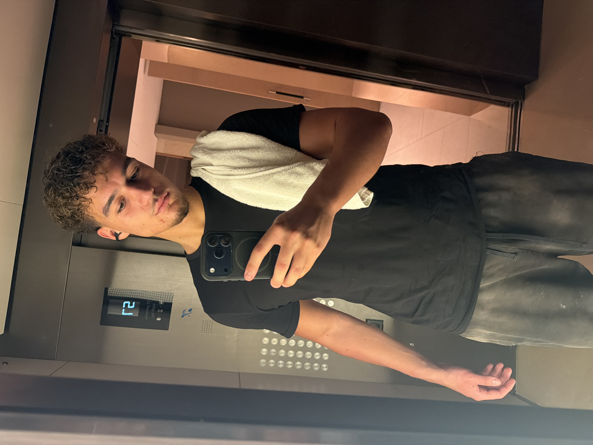 A male is taking a selfie in what appears to be an elevator. He has curly hair, a towel draped over his shoulder, and is holding a phone in his hand. The elevator panel shows the number "21" and some accessibility symbols. The background visible through the elevator doors suggests an interior space, possibly a hotel or residential building. There is no explicit evidence in the image to confirm the location as Dubai, United Arab Emirates.