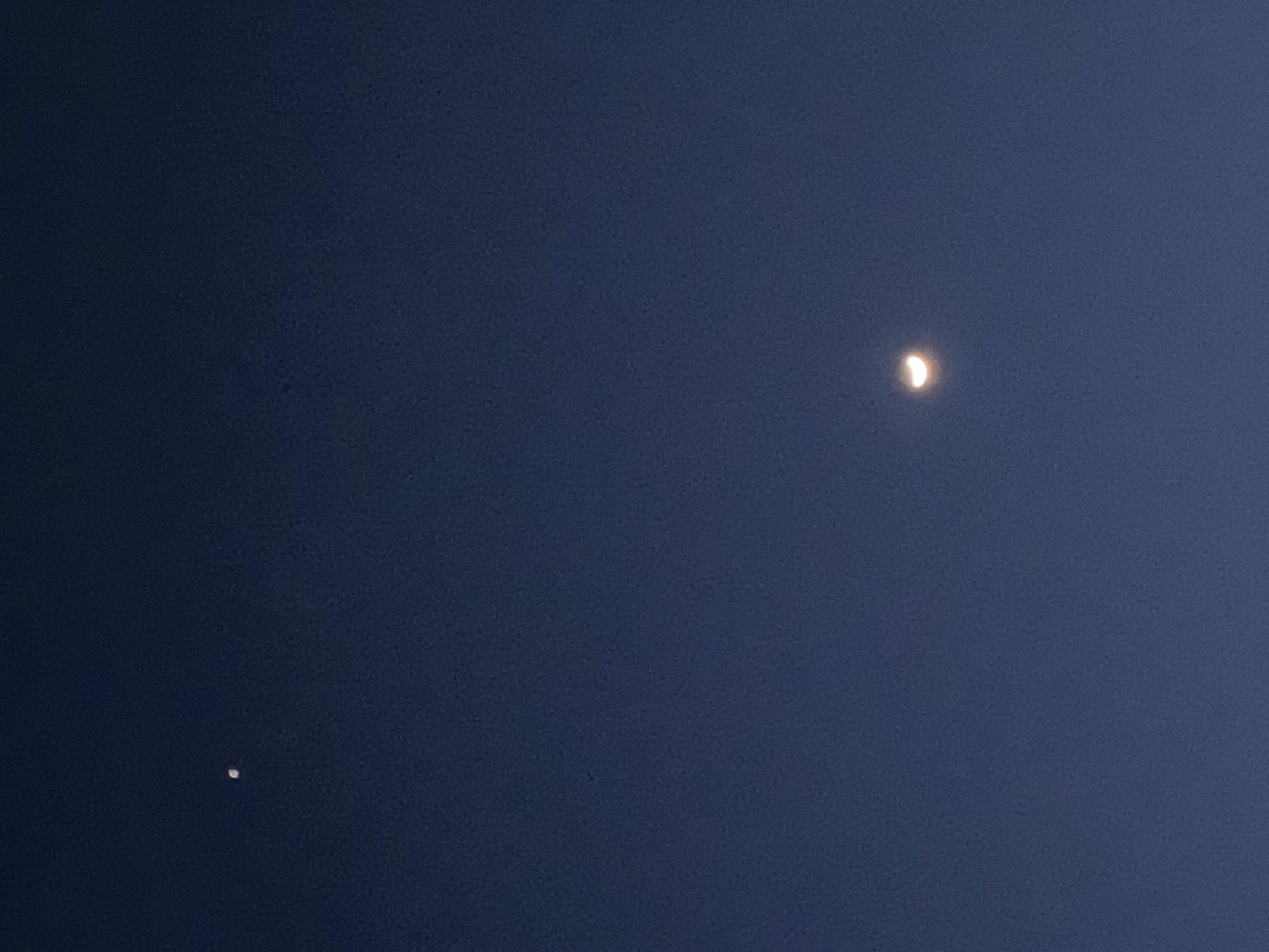 The image captures a serene twilight or nighttime sky over Alanya, Turkey. The deep, uniform dark blue hue of the sky suggests a period just after sunset or before sunrise, or a clear night with some ambient light. Two prominent celestial bodies are visible against this backdrop. In the upper right portion of the frame, a distinct crescent moon shines brightly, with its illuminated curve facing towards the left. Below and to the left of the moon, in the lower-left quadrant, a smaller, intensely bright, punctiform object is visible, resembling a planet or a very bright star. The sky is notably clear, with no clouds or atmospheric obstructions, allowing for an unobstructed view of these celestial features. There are no people, man-made structures, or visible text within the photograph. The scene is one of quiet astronomical observation.