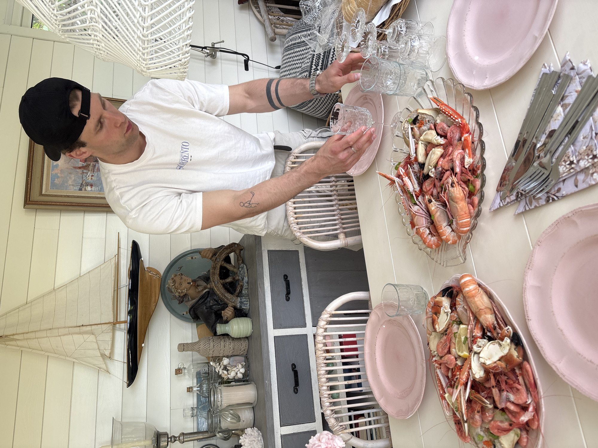 In Larkollen, Norway, a man wearing a black baseball cap and a white t-shirt with the word "SORRENTO" printed on it is setting a table on an outdoor patio. He is placing a glass on a pink plate. The table is laden with seafood, including langoustines and shrimp, served in clear glass dishes. There are also pink plates, cutlery, and wine glasses. In the background, a nautical-themed decor is visible, including a model sailboat and a figurine of a man at a ship's wheel. The patio appears to be an extension of a house, with wooden plank walls and a white roof.