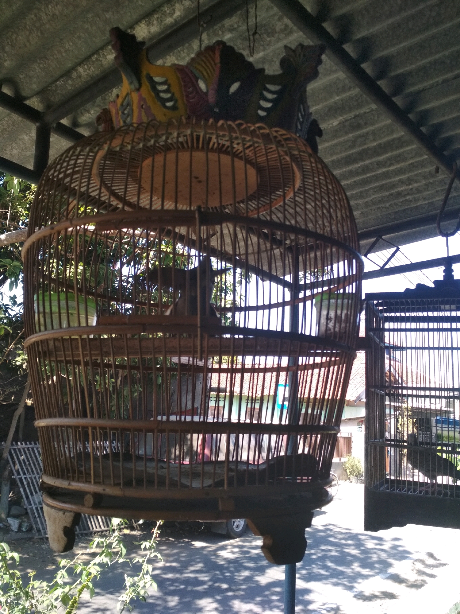 A large, ornate, round birdcage made of bamboo is suspended from a metal roof structure in what appears to be a village setting. The cage is intricately woven and features a decorative wooden element at the top, resembling carved birds. Inside the cage, there are wooden perches and a green object, possibly a water or food container. To the right, a second, smaller, dark-colored metal birdcage is also visible, containing a bird. The background shows trees, vegetation, and glimpses of buildings. The scene is illuminated by sunlight, casting shadows on the ground, which is paved and shows some gravel. The location is identified as Desa Pamijen, Kecamatan Sokaraja, Indonesia.