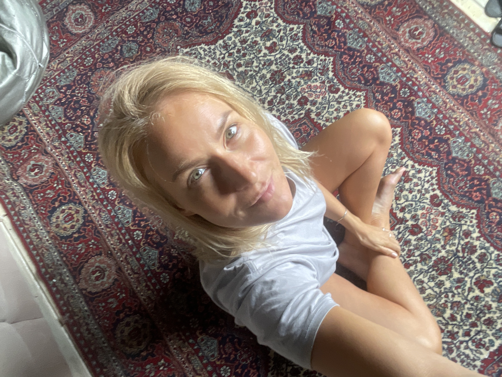 A woman with blonde hair and fair skin is seated on an ornate rug. She is wearing a light grey t-shirt. Her legs are crossed and she is holding one hand over the other. The rug has a complex pattern of red, blue, beige and brown colours, and small, repeating motifs. A metallic beanbag chair is visible in the upper-left corner. The image appears to have been taken from an overhead perspective.
There is no information in this image to allow an accurate description of Baranovka, Russia.