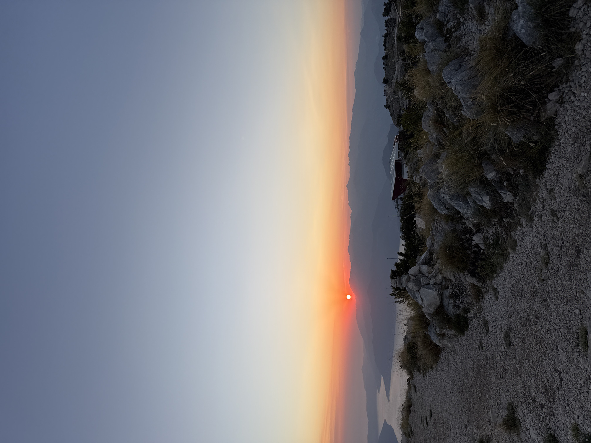 The image captures a sunset view from a mountain in Kuk, Montenegro. The sun, a bright orange disc, is low on the horizon, casting a warm glow across the sky and illuminating the distant mountains and the body of water in the foreground. The terrain in the foreground consists of a gravel path and rocky outcrops covered with dry grass. In the middle ground, there are some structures, including a building with a red roof and several smaller buildings, as well as a few people gathered near them. The background is dominated by layers of silhouetted mountains fading into the hazy distance. The sky transitions from a pale blue at the top to a warm orange and pink near the horizon.