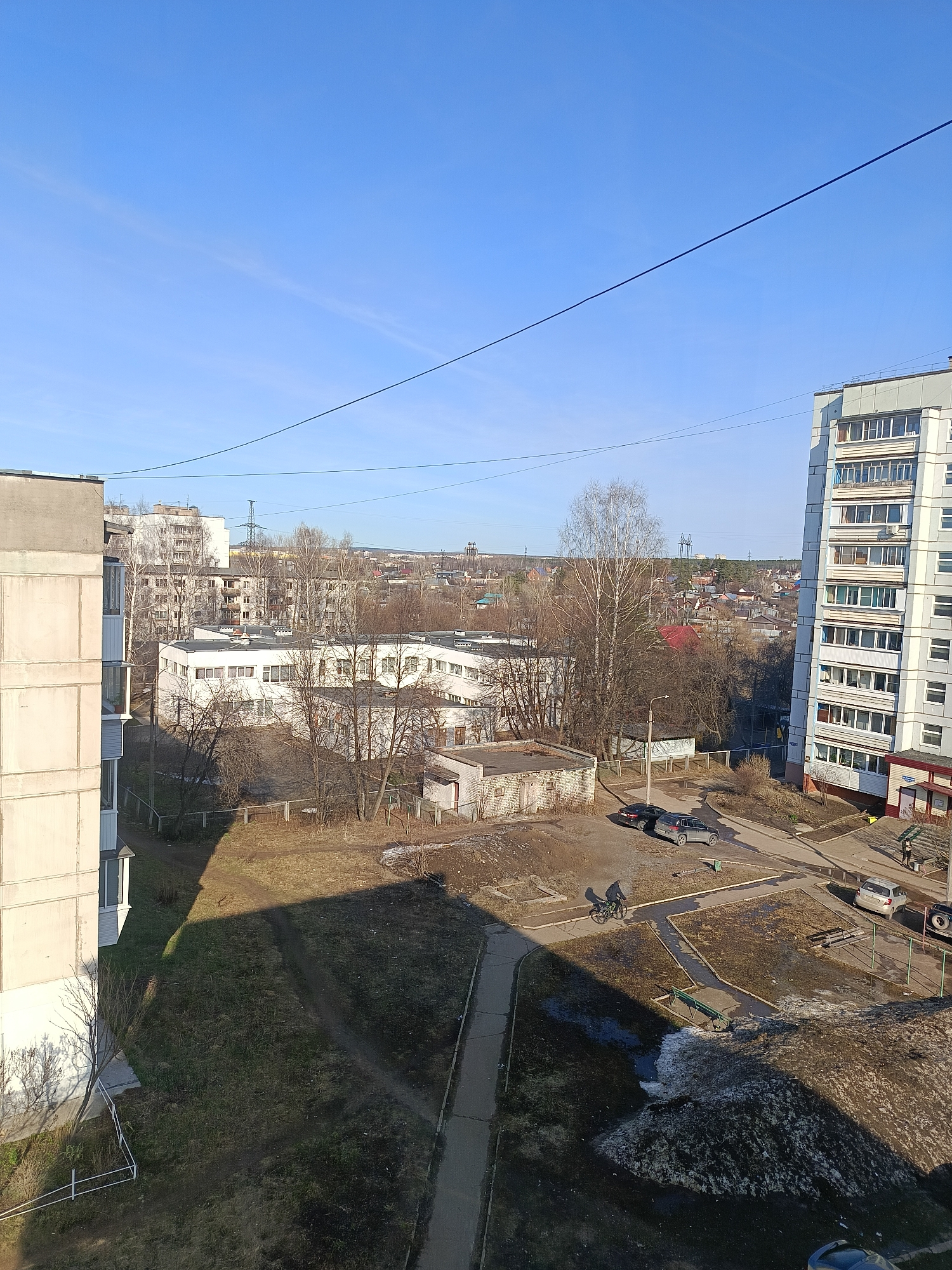 The image is taken from an elevated position, likely from an apartment building, overlooking an urban landscape in what appears to be the city of Chaikovsky, Russia. The sky is clear and blue. Several buildings are visible, including multi-story apartment blocks and what appears to be a lower-rise building, possibly a school or administrative structure. A person is riding a bicycle on a paved walkway in a courtyard area. There are a few cars parked nearby. Patches of melting snow are observed in the yard and surrounding areas, indicating it is likely early spring. Bare trees are scattered throughout the scene, and power lines stretch across the top of the frame.