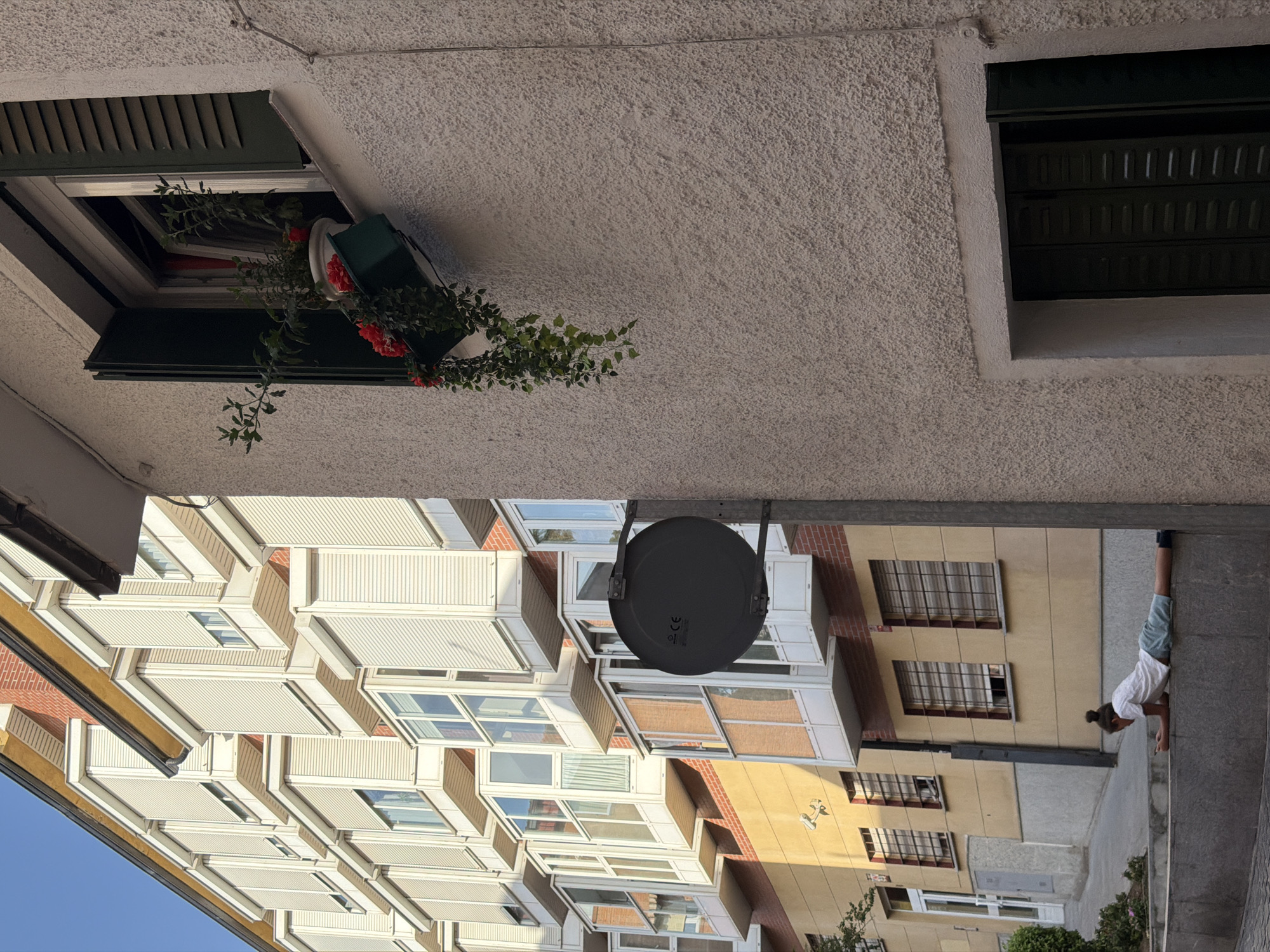 The image captures a street scene in Las Rozas de Madrid, Spain, on a sunny day. In the foreground, a young person is lying face down on a stone ledge, holding a smartphone. They are wearing a white t-shirt and light blue shorts. Behind them, a beige building with yellow and brick accents features several windows with roller shutters and bay windows with glass. A dark circular satellite dish is mounted on the side of this building. To the left, another building with white bay windows and beige and brick facades is visible, with a clear blue sky above. A planter box with red flowers and trailing green plants hangs from an open window on a lighter-colored building on the right. The overall environment suggests an urban residential area.