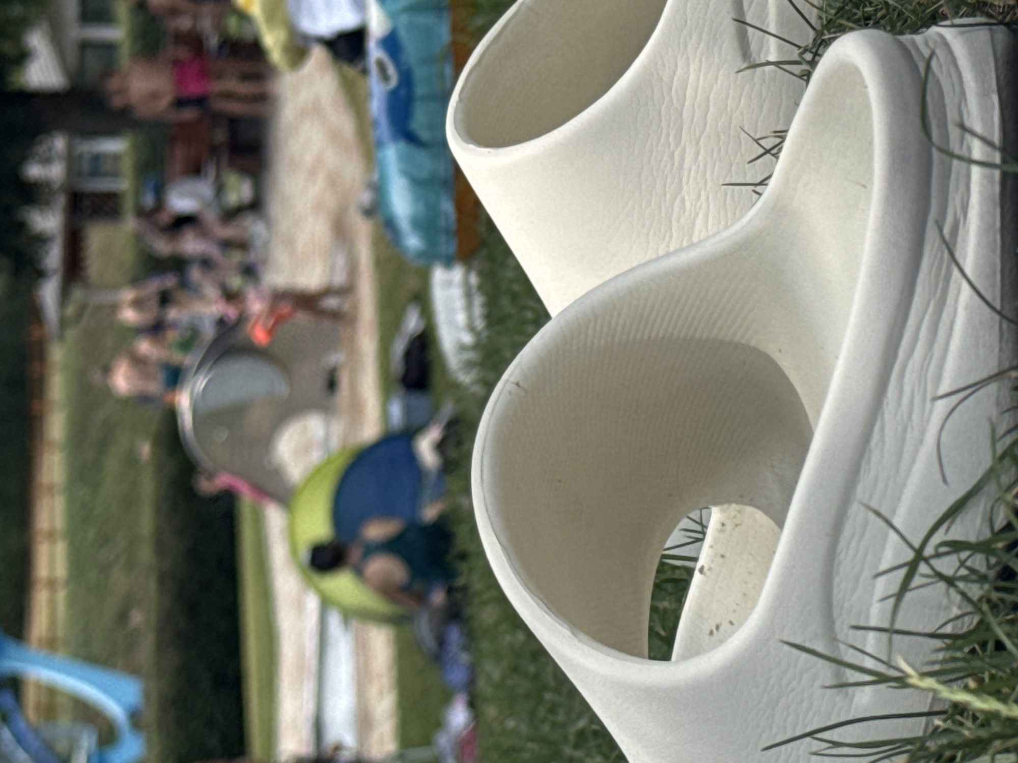 This image captures a scene at the Hainburg an der Donau outdoor swimming pool area in Austria. In the foreground, a pair of light-colored slides are placed on grass. In the mid-ground, several people are visible in the background, some near a tent and others in a gathering. There is also a blue and orange inflatable tube visible. The overall environment suggests a recreational setting with a focus on outdoor activities.
