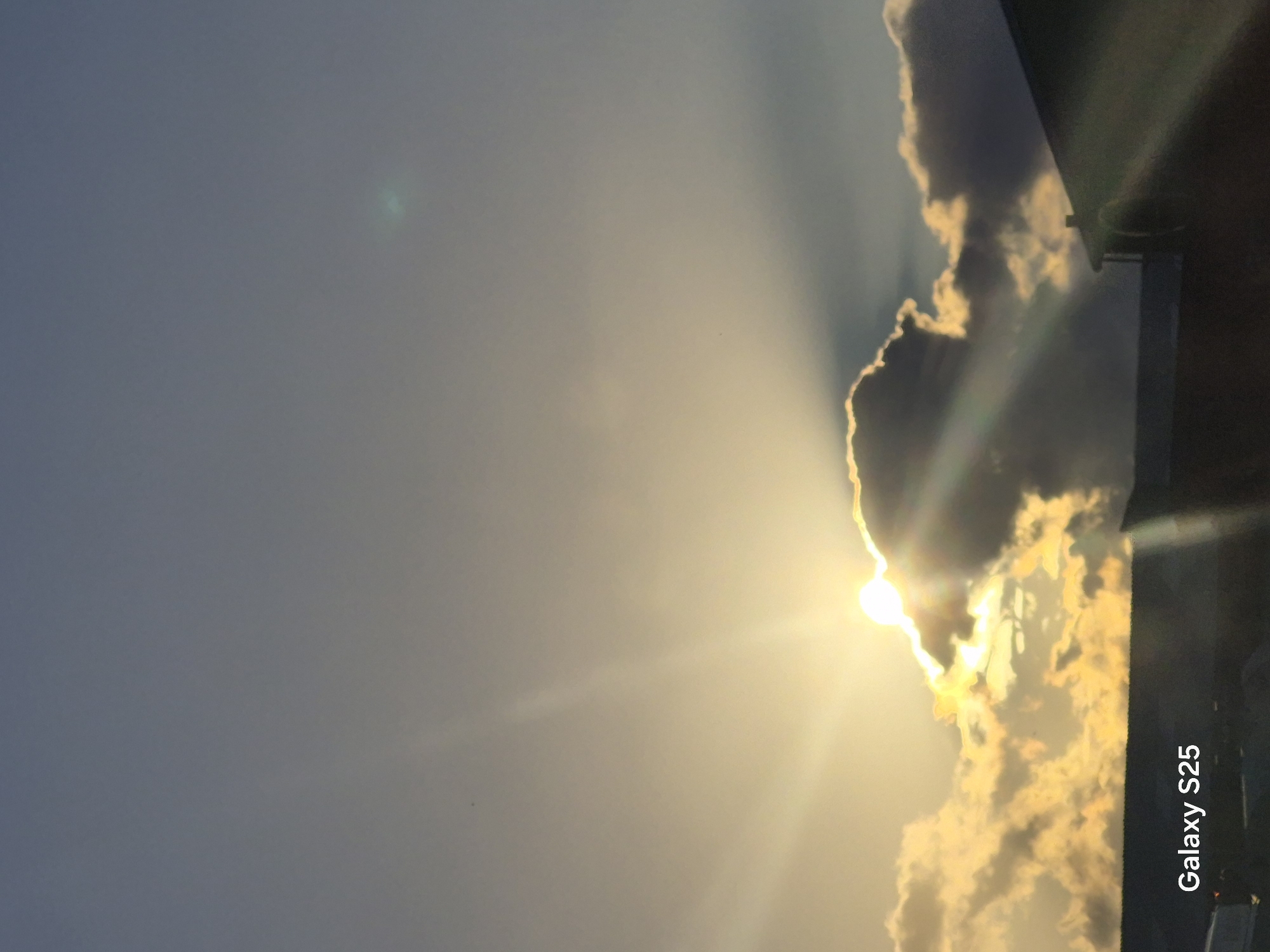 The image captures a dramatic sunset over Pątnów, Poland. The sun, a brilliant orb partially obscured by dark, heavy clouds, casts long, ethereal rays of light across the sky. These crepuscular rays are illuminated by the warm, golden hues of the setting sun, highlighting the edges of the clouds and creating a stark contrast with the darker, shadowed portions. In the foreground, the silhouette of rooftops suggests an urban or suburban environment. The lower left corner of the image contains the text "Galaxy S25," indicating the device used to capture the photograph. The sky transitions from a light, hazy blue at the top to a warmer, more intense yellow and orange near the horizon.
