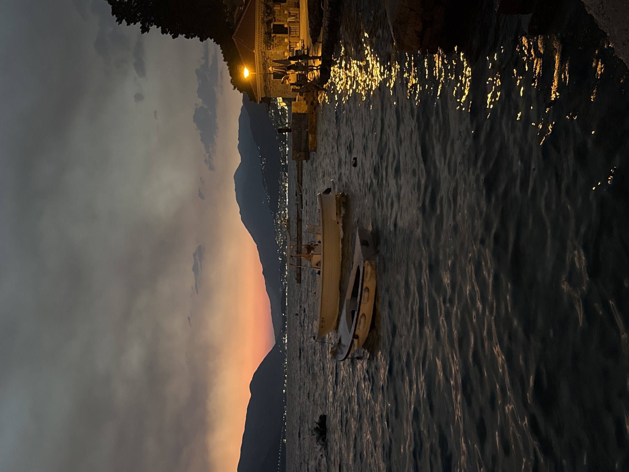 This is a vertical, eye-level, medium shot of a bay at dusk, with the water in the foreground and mountains and a building in the background. The sky is dark gray with streaks of orange and pink near the horizon. The water is dark blue and choppy, with reflections of a light from a building on the right side of the frame. There are three boats in the water; two small white rowboats in the foreground and a larger white boat in the mid-ground. On the right side of the frame, a building with a red tile roof is lit by a single yellow outdoor light. Three people are standing on the stone embankment in front of the building. The mountains in the background are dark and silhouetted against the sky.