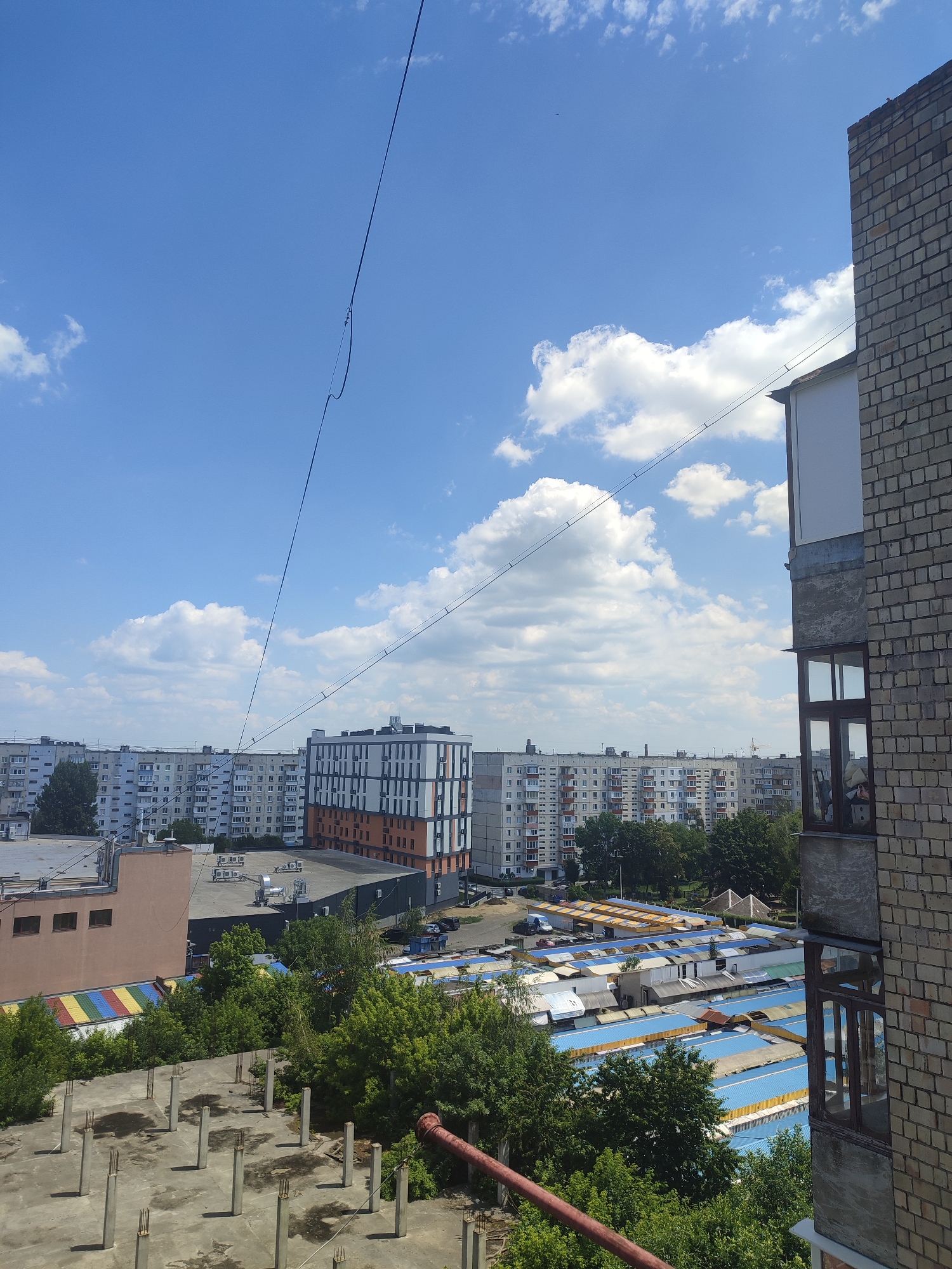 The image depicts a scene of urban residential buildings and a marketplace in Chernivtsi, Ukraine. In the foreground, a partially constructed concrete building with rebar-reinforced columns is visible, suggesting ongoing or abandoned construction. To the right, a brick building with windows occupies the edge of the frame. The middle ground features a series of low-lying market stalls with blue and yellow awnings, characteristic of an outdoor market. Behind the market, a row of uniform, multi-story apartment buildings stretches across the horizon. A more modern building with a white, orange, and dark-gray facade stands out amongst the older structures. The sky is a bright blue with scattered white clouds. The overall impression is a snapshot of everyday life and development within a Ukrainian city.