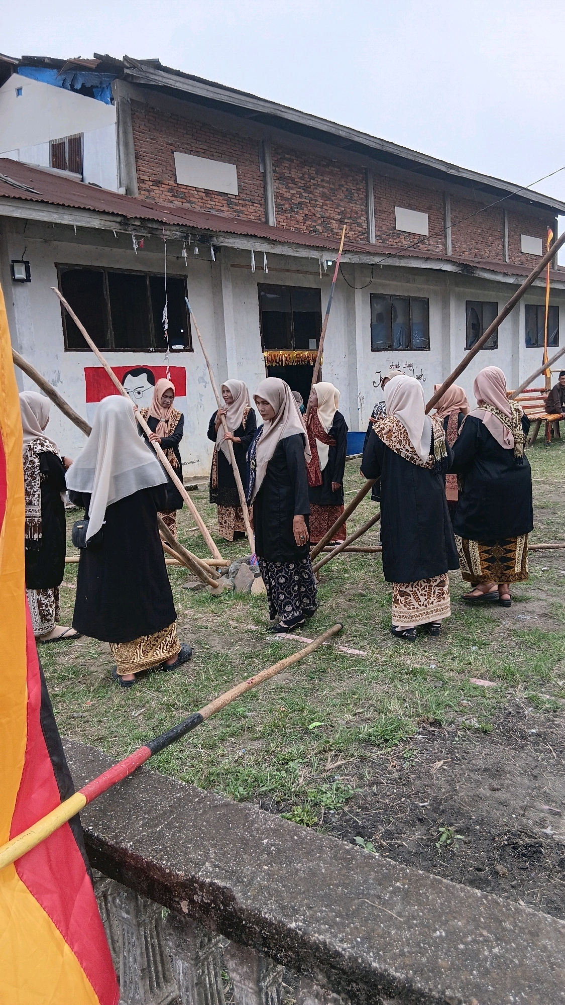 The image depicts an outdoor scene in Padang Laweh, Indonesia, where a group of eight women, all wearing traditional Islamic attire including hijabs and long dresses, are gathered around a structure made of wooden poles. The women are positioned in various stances, some holding or interacting with the poles, suggesting they might be involved in setting up or dismantling the structure, or perhaps participating in a ceremony or activity related to it. They are dressed in dark-colored outer garments and patterned skirts. The background features a weathered building with a corrugated metal roof and visible brickwork. The ground is covered with grass and dirt. In the foreground, a concrete barrier with decorative carvings is visible, and a flag with red, yellow, and black colors is partially displayed. The overall atmosphere appears to be a community gathering or event.