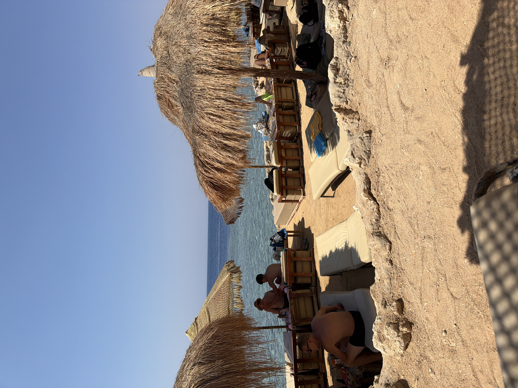 This image depicts a beachfront scene in Sharm El Sheikh, Egypt. In the foreground, the sandy beach is occupied by several lounge chairs and low seating areas, some covered by straw umbrellas. Some of these seating areas have cushions and are set up for relaxation. A man with sunglasses is seated in a lounge chair in the lower left portion of the frame, facing away from the camera. Further back, under a straw umbrella, two men are seated and appear to be interacting, with one placing his hand on the other's shoulder. The clear blue sea stretches out behind the beach, meeting a cloudless sky. The overall atmosphere suggests a tranquil day at a resort or beach club.