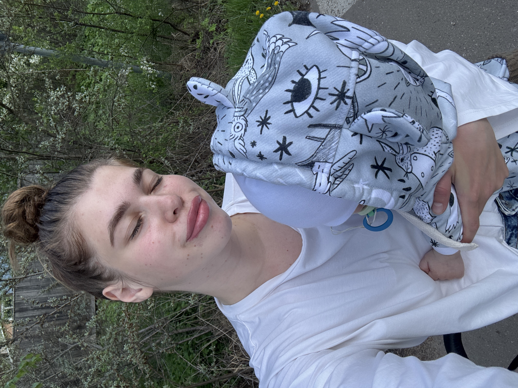 A woman with her eyes closed and hair in a bun holds a baby wearing a patterned hat with animal designs and a pacifier attached. The location appears to be outdoors near trees and foliage.