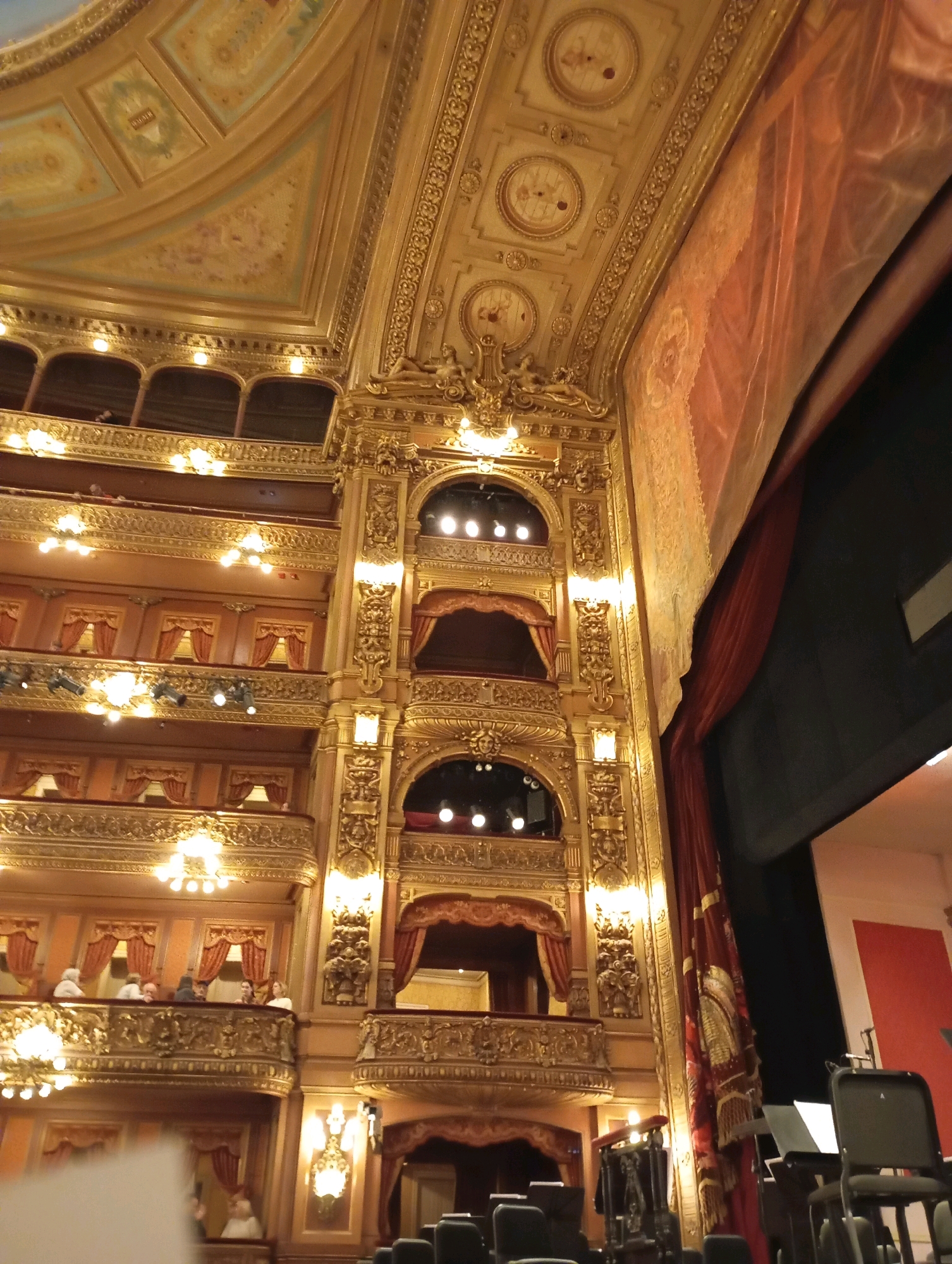 This image shows the interior of the Teatro Colón in Buenos Aires, Argentina, a renowned opera house. The ornate, gilded architecture is prominently featured, with multiple tiers of balconies adorned with intricate carvings and golden detailing. Large, decorative chandeliers hang in some of the boxes, and small lamps illuminate the balconies. The ceiling is also elaborately decorated with painted panels and gilded trim. A large red curtain hangs on the right side of the frame, partially obscuring the stage area. Some audience members are visible in the balconies, seated and looking out. The overall impression is one of opulence and historical grandeur.