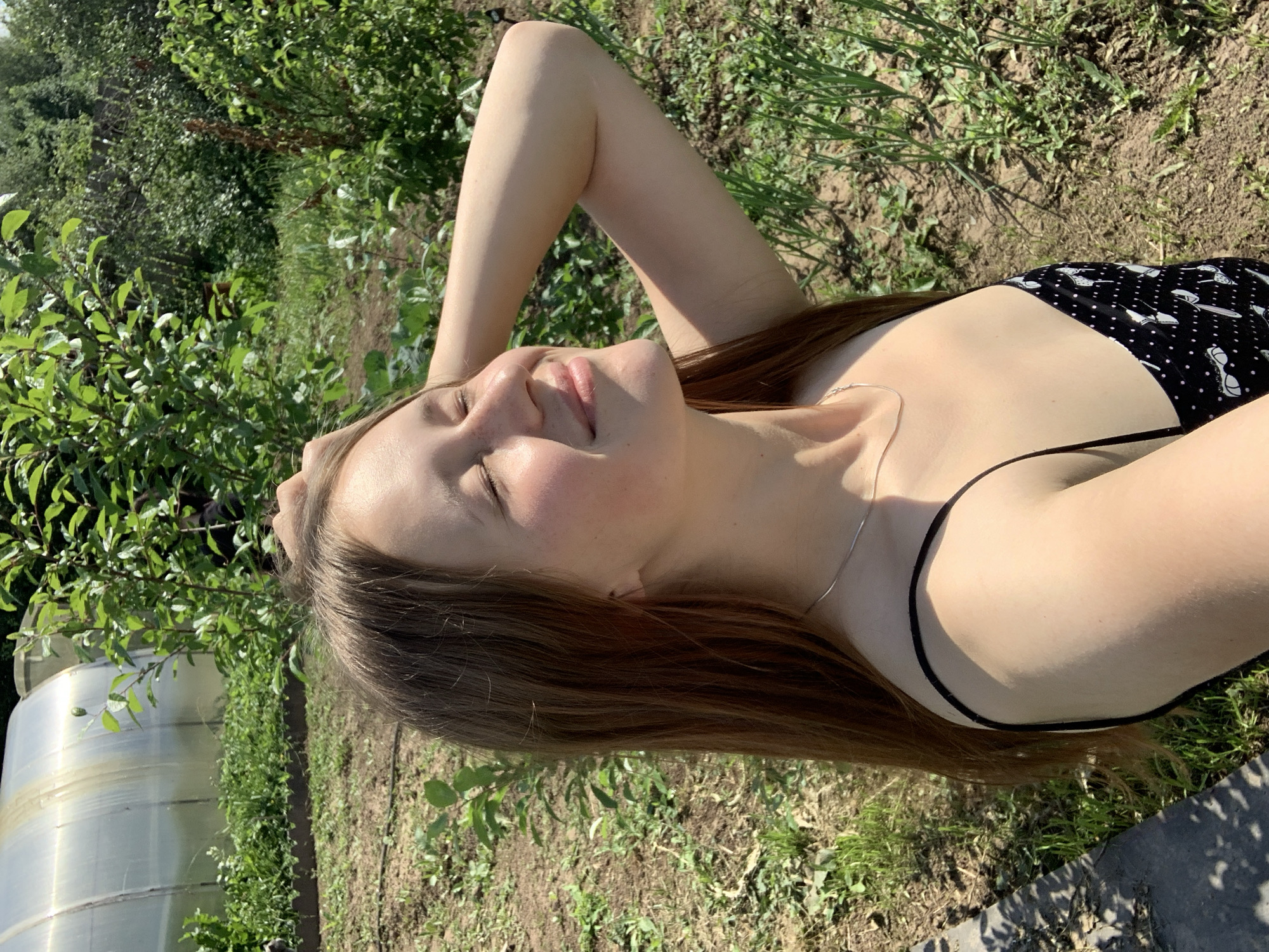 A young woman is lying on the ground outdoors, with her head propped up on her arm. She is wearing a black bikini with a white polka dot pattern. Her eyes are closed and she has a serene expression on her face, with her mouth slightly open. Sunlight is shining on her face, highlighting her features. Her long, brown hair is spread out around her. In the background, there are lush green trees and plants, and a greenhouse structure is visible on the left side of the image. The ground appears to be made of dirt and grass. The lighting suggests it is daytime, likely a sunny day. The overall mood is relaxed and peaceful. No text is visible in the image.