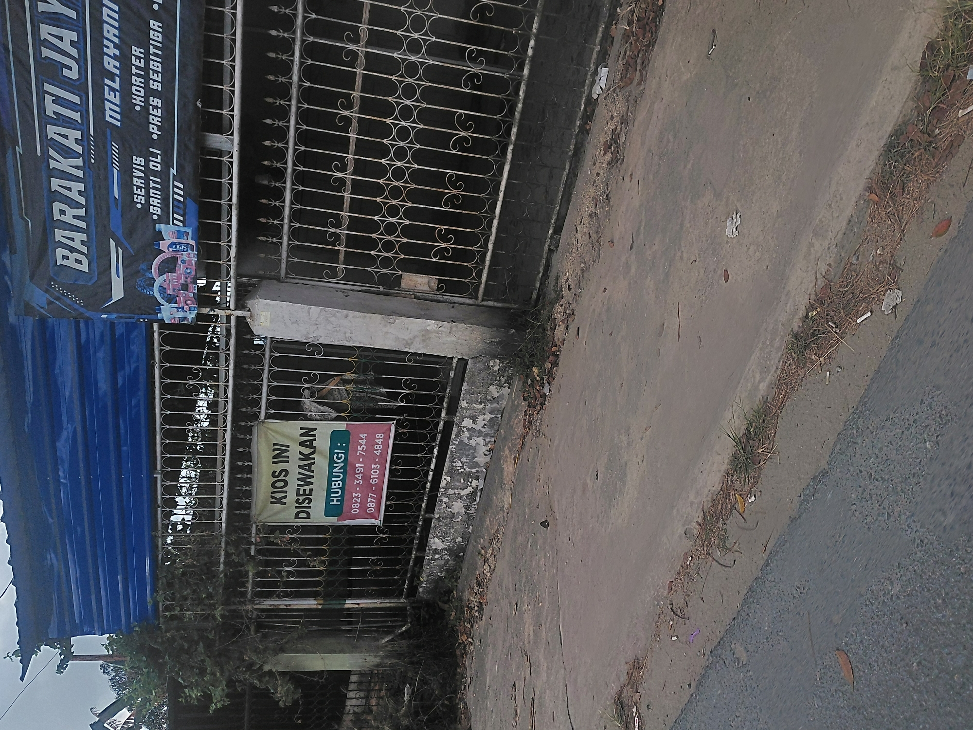 The image depicts a street scene in Kendari, Indonesia. On the left, a narrow road runs diagonally, bordered by a gray concrete sidewalk littered with dried leaves and debris. A dark gray asphalt road is visible to the left of the sidewalk.
Dominating the right side of the image is a metal gate, intricately designed with swirling patterns. Behind the gate, a building with a blue corrugated metal roof is partially visible. A banner is attached to the gate, displaying text in Indonesian. The top section of the banner reads "BARAKATI JAY" in large, stylized white lettering against a dark background. Below this, smaller text lists services: "MELAYANI" (serving) followed by bullet points: "• SERVIS" (service), "• KORTER" (overhaul), "• GANTI OLI" (oil change), and "• PRES SEGITIGA" (triangle press).
Further to the left on the gate, a yellow sign is prominently displayed. It reads "KIOS INI DI SEWAKAN" (This kiosk is for rent) in black, bold lettering. Beneath this, in a smaller font, it says "HUBUNGI:" (Contact:) followed by two phone numbers: "0823 - 3491 - 7544" and "0877 - 6103 - 4848".
Vegetation, including a dark, leafy tree, grows to the left of the gate, partially obscuring the building. The overall environment appears to be an urban street with commercial activity indicated by the signage. The lighting suggests it is daytime, possibly overcast.