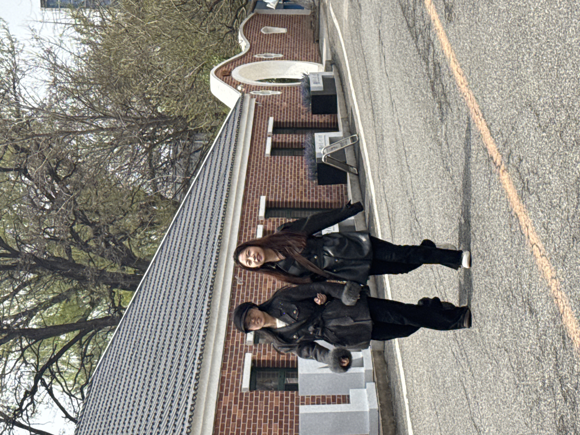 Two women are standing outdoors, posing for a photograph. The image, oriented correctly, shows them positioned in front of a building with a road to their right.
The woman in the foreground is wearing a dark, possibly wool or thick fabric, jacket or coat, dark pants, and a dark cap. Her hands are visible, wearing dark gloves or having fur-trimmed cuffs. She has dark hair and a neutral expression, looking directly at the camera.
Behind her, the second woman is dressed in a dark, shiny material jacket (possibly leather or faux leather), dark pants, and white sneakers. She has long, dark hair and a slight smile while looking towards the camera.
They are standing on a paved surface next to a building characterized by red brick walls and a dark gray, perhaps tiled, roof. The building features distinctive white architectural elements, including a prominent arched opening and other decorative trim. Several small rectangular windows are visible in the brick facade. In front of the building, there are light-colored stone or concrete structures, possibly planters or steps.
To the right of the women and the building is an asphalt road with a faded orange or yellow lane line and a white edge line. In the background and to the left of the building, bare deciduous trees with numerous branches indicate a cooler season, such as late autumn or winter. The scene is illuminated by bright, natural daylight. No discernible text is visible anywhere in the image. The overall setting appears to be an urban or semi-urban environment.