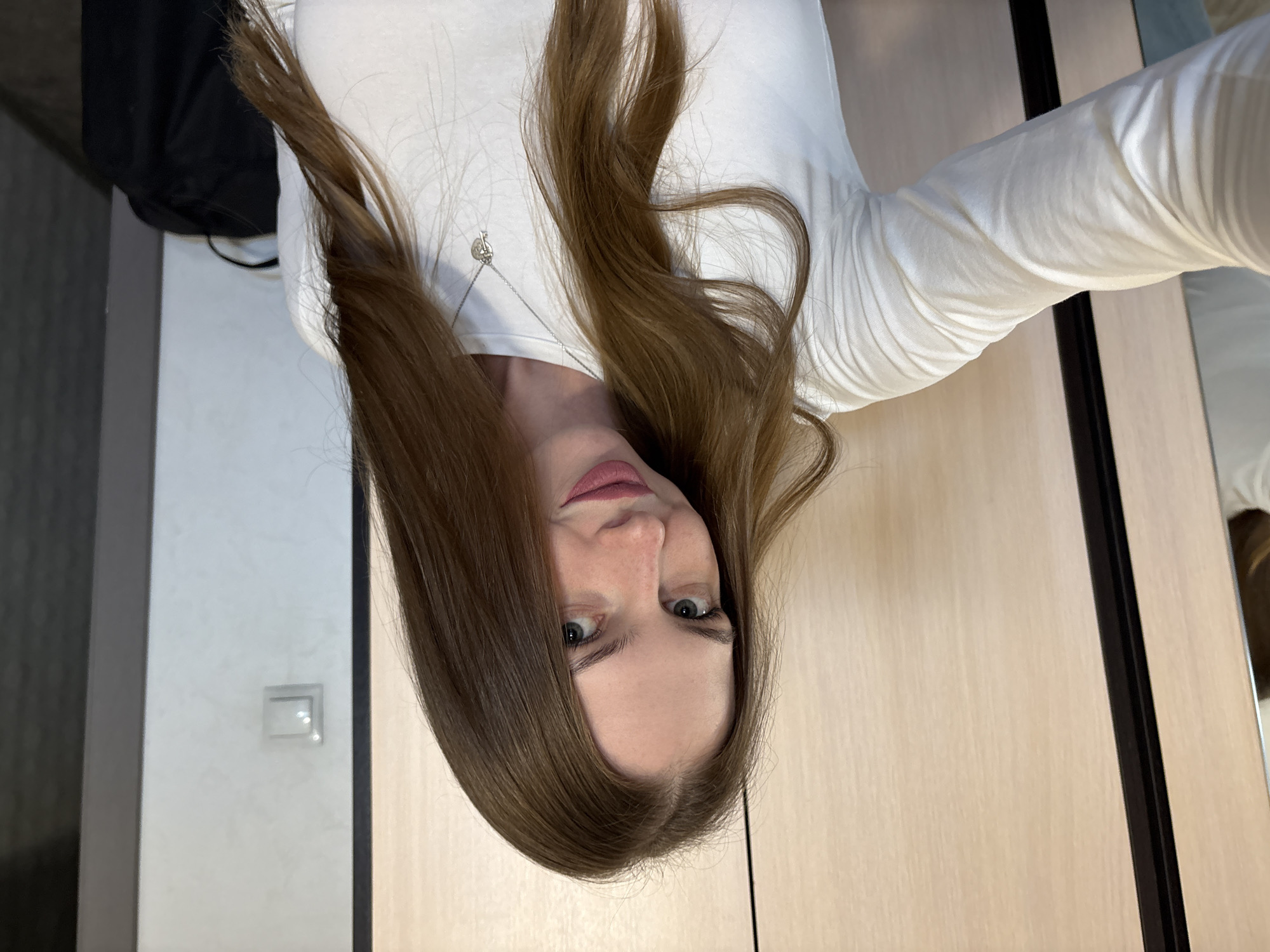This is a vertical, head-on selfie of a young woman with long, brown hair and blue eyes. She is wearing a white long-sleeved shirt and a silver necklace with a small, key-shaped pendant. Her hair is parted in the middle and falls loosely around her face. She is looking directly at the camera with a neutral expression. The background appears to be an indoor setting, possibly a room with light-colored wooden furniture. The lighting is soft and natural, suggesting it might be daytime. There are no visible text elements in the image. The overall impression is one of a casual, everyday moment captured in a personal photograph.