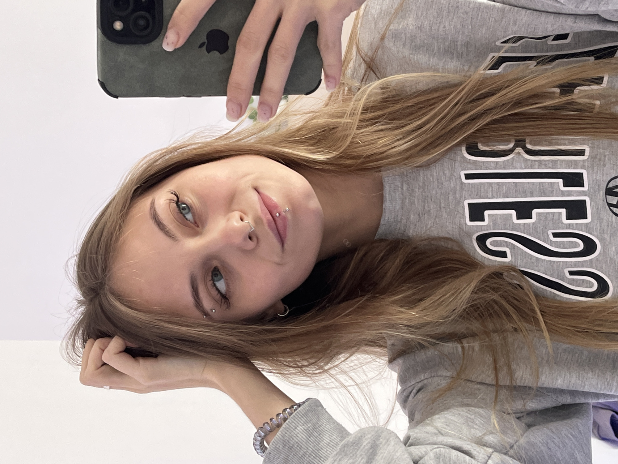 A young woman with long, light brown hair is holding a phone in her left hand to take a selfie. She is wearing a gray sweatshirt with large black letters and a small black design visible on the right side of the image. She has several piercings: one in her eyebrow, one in her nose, and two in her lip. She is wearing a bracelet on her left wrist. The background appears to be a plain white wall. The image does not provide any information about the location in the city of Фоки, Russia.