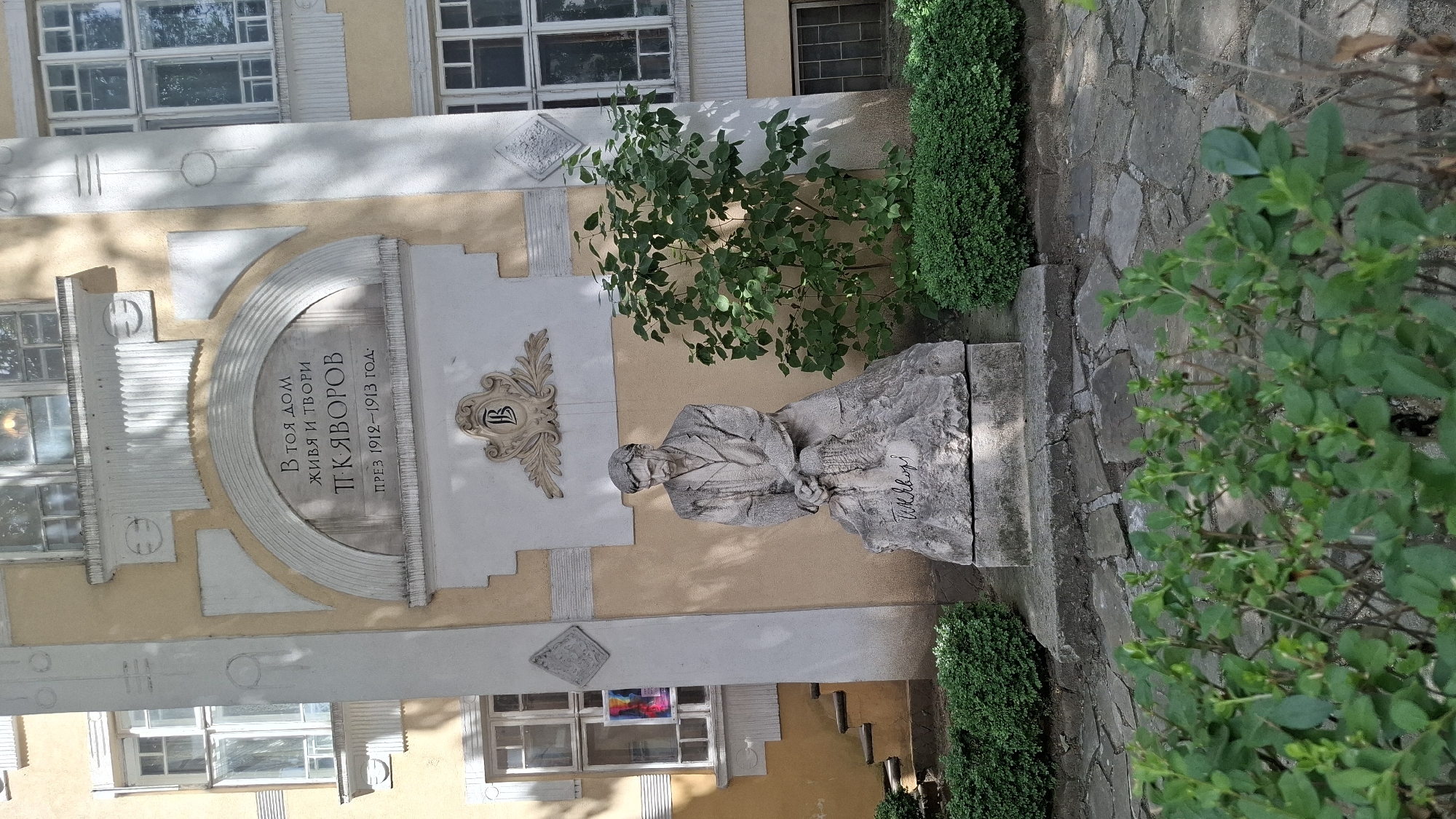The image shows a stone sculpture of a man seated on a rock, with his hands clasped in front of him. He is wearing a coat and appears to be looking down. Above the sculpture, on the wall of a building, is a plaque with Cyrillic writing and a decorative emblem. The building has windows and is painted yellow and cream. There are also hedges and bushes in front of the building and around the sculpture. The ground is paved with irregular stones. Based on the plaque which reads "В ТОЯ ДОМ ЖИВЯ И ТВОРИ П.К. ЯВОРОВ ПРЕЗ 1912-1913 ГОД.", this is the Peyo Yavorov House-Museum, located in Sofia, Bulgaria. The plaque commemorates that the poet Peyo Yavorov lived and worked here in 1912-1913.