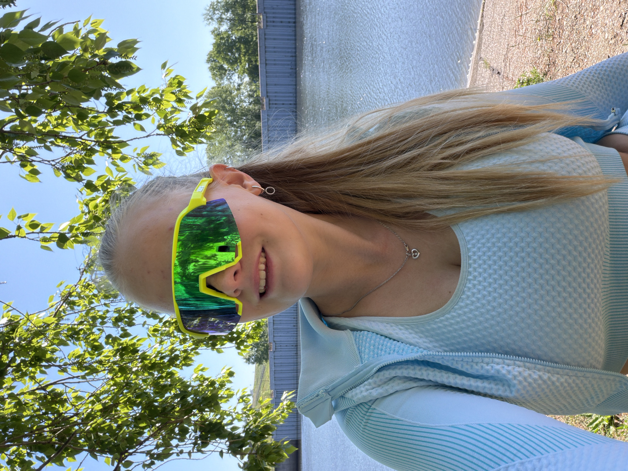 A woman is photographed outdoors near the city of Chaikovsky, Russia. She is wearing lime green sunglasses with reflective green lenses, a light blue textured top, and a zip-up jacket. She has long blonde hair and is wearing a silver necklace and hoop earrings. The background includes a body of water and trees.