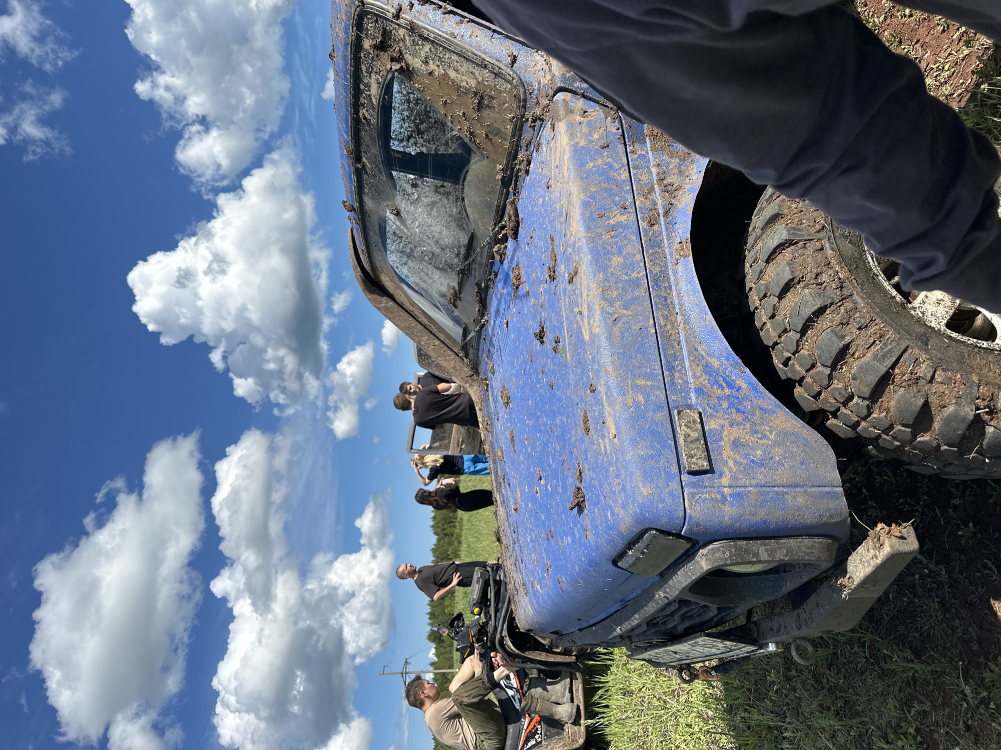 A heavily mud-splattered, blue off-road vehicle is the primary subject of the image. It appears to have been involved in off-road activities given the extent of mud coverage. A group of people stands near the vehicle, possibly inspecting it or preparing to assist. The setting is a grassy field with a clear blue sky dotted with fluffy white clouds. Based on context from surrounding images, the location is in or near Ilyinsky, Russia.