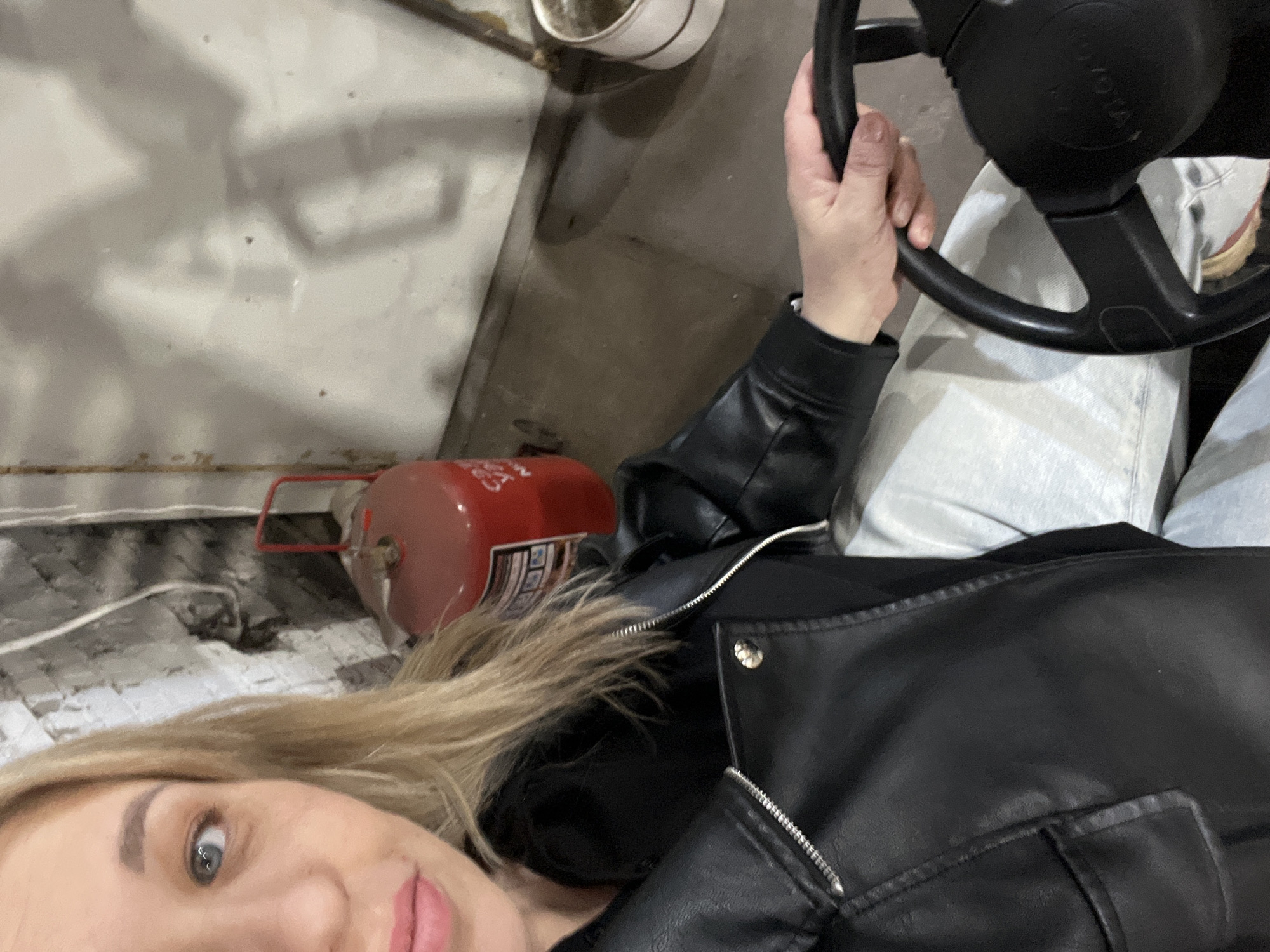 A woman takes a selfie from a diagonal perspective inside what appears to be a vehicle or an industrial setting. Her face, with blonde hair, a wide blue eye, and pink lipstick, is visible in the lower-left corner, looking towards the camera. She is wearing a black leather jacket with a zippered front and a buttoned lapel, over light-colored trousers or jeans.
Her left hand rests on a black steering wheel, which dominates the upper-right portion of the frame. Faint, unreadable text, possibly "TOYOTA," is visible on the center of the steering wheel. To her left, lying on its side, is a red cylindrical object resembling a fire extinguisher or a small propane tank, featuring a barcode and indistinct text on a label. A white cup or container is partially visible in the upper-middle background.
The surroundings suggest an indoor, utilitarian environment, possibly a garage or workshop, with a light-colored, textured wall or surface on the left and a darker, less defined surface on the right. The lighting is even, characteristic of indoor artificial light. The woman displays a neutral to slightly smiling expression. The location context is Chaikovsky, Russia.