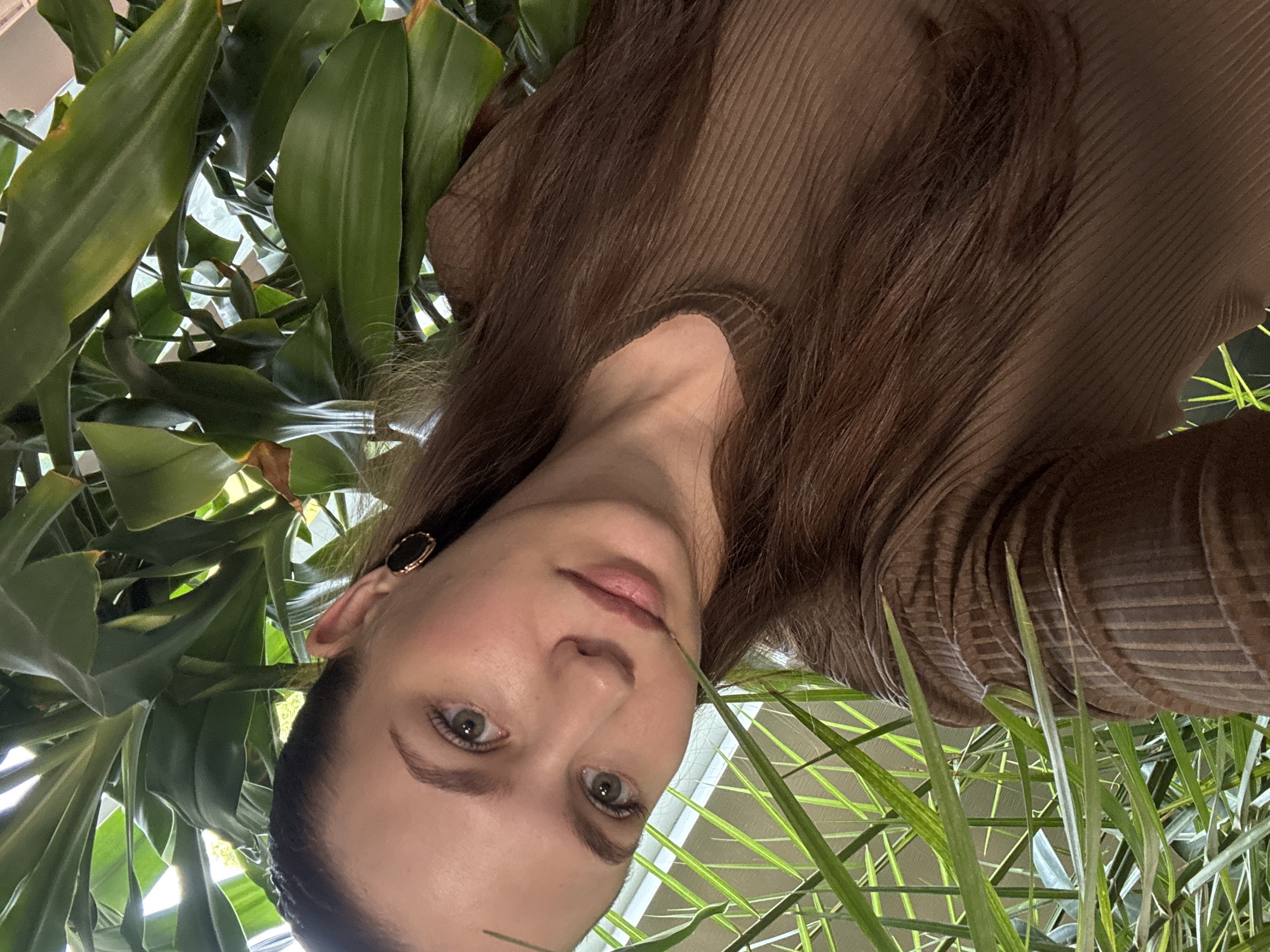 A medium close-up portrait features a young woman looking directly towards the camera, surrounded by lush green foliage. The image is oriented 90 degrees counter-clockwise. The woman has long, straight, dark brown hair, fair skin, and light-colored eyes, appearing green or hazel. Her lips are naturally full with a soft pink hue, and her expression is neutral and calm. She is wearing a brown, ribbed, long-sleeved top with a simple neckline. A distinctive black and gold earring adorns her visible left ear.
The setting is dominated by numerous large, broad-leafed green plants that frame her face and body, creating a dense, verdant backdrop and foreground. The leaves vary in shade from vibrant to darker greens, with some showing lighter accents from the natural light. Bright, diffuse light illuminates the scene, suggesting an indoor environment such as a conservatory, greenhouse, or a room with many large houseplants, possibly near a window, rather than an outdoor setting given the location context of Nizhnyaya Gar', Russia. No specific time of day or weather conditions are visible beyond the clear natural light. There is no visible text in the media.