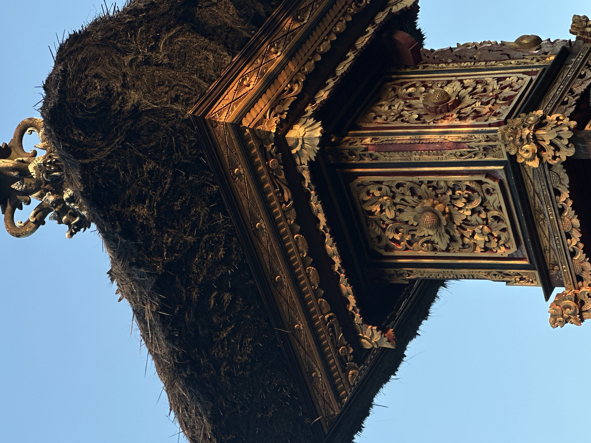 The image shows a detailed view of a traditional Balinese structure in North Kuta, Indonesia. The structure features an elaborately carved wooden facade with gold detailing. The carvings depict floral and foliate motifs. Above the wooden structure is a thatched roof made of dark, coarse natural fibers. The background is a clear, light blue sky. The perspective is from below, angled upwards towards the structure.