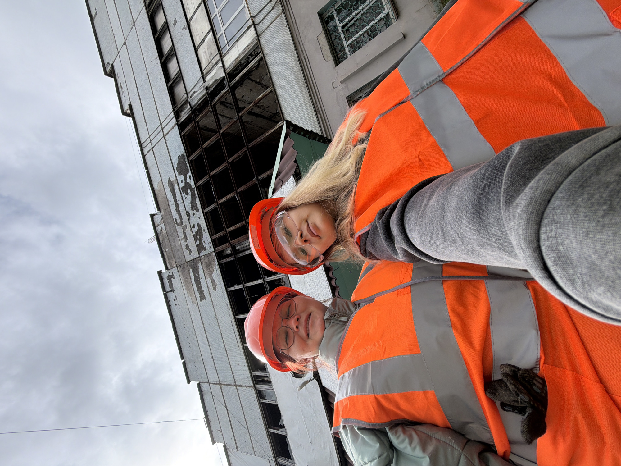 This low-angle, tilted photograph captures two individuals, likely young women, posing for the camera with a dilapidated building and an overcast sky in the background. The scene suggests a construction, demolition, or inspection environment.
Both individuals are wearing bright orange high-visibility vests with grey reflective stripes and safety hard hats. The person closer to the camera has long blonde hair, safety glasses, and a reddish-orange hard hat, looking upwards with a playful expression, tongue slightly out. The second person, positioned behind and slightly below the first, wears a similar orange hard hat and prescription glasses, smiling broadly. Black gloves are visible tucked into the pocket of one of the vests.
The background features a multi-story building with a somewhat industrial or utilitarian architectural style. Its facade is light-colored, possibly made of panels or tiles, showing significant signs of wear, damage, and staining. Many windows are broken or missing, revealing dark, empty interiors or structural elements. The sky is uniformly overcast, indicating a cloudy daytime. The image's severe tilt makes the building appear to lean dramatically. No visible text is present in the image. Given the location context, this scene is set in Chaikovsky, Russia.