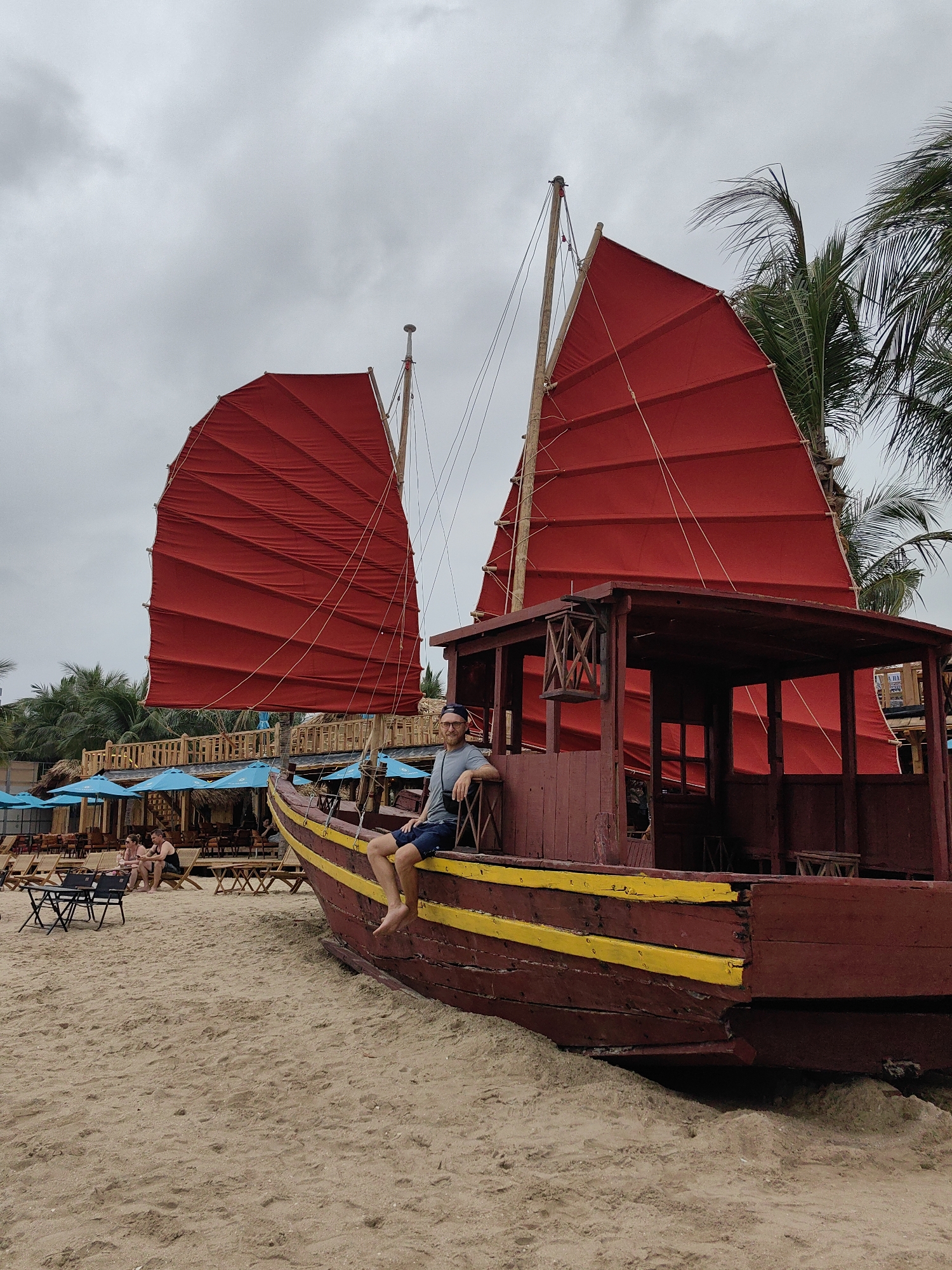 A man sits on the side of a grounded, brown, junk boat on a sandy beach. The boat has two red sails and two yellow stripes along the hull. The boat is located on Bai Chay Ward, Vietnam. A few people are seated in chairs under blue umbrellas in the background. Palm trees are visible in the background as well. The sky is overcast.