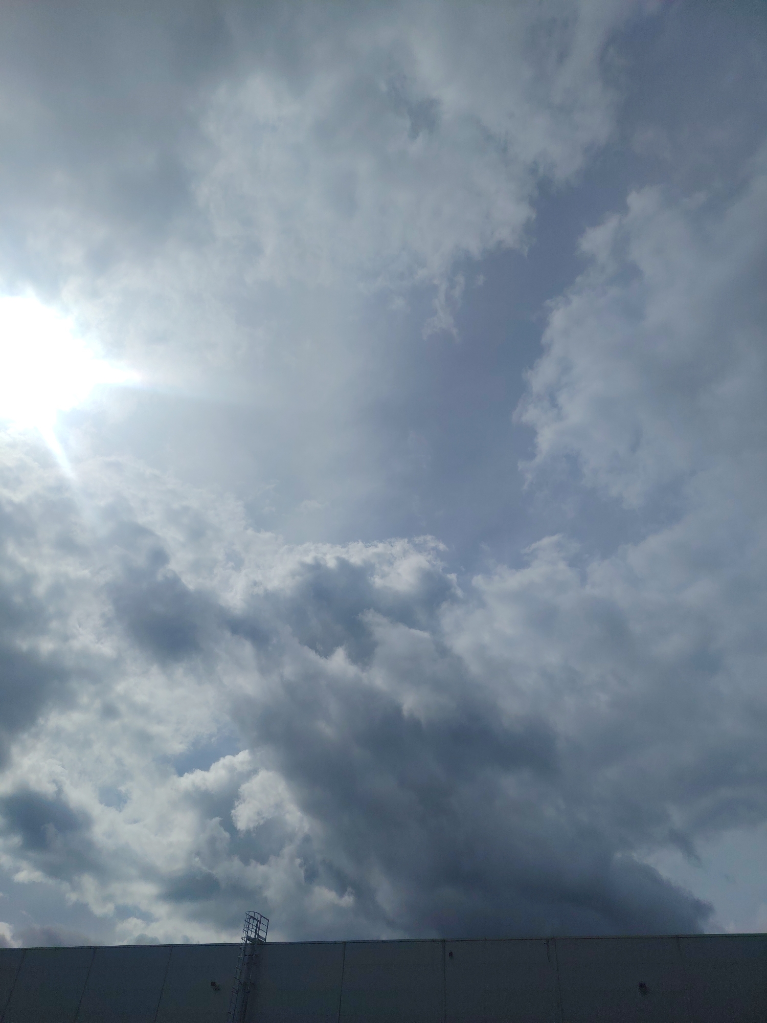 The image shows a partially cloudy sky with the sun visible on the left, casting a bright glare. The clouds are a mix of light and dark gray, with some appearing fluffy and others more dense. In the foreground, the top of a light-colored building is visible, extending across the bottom of the frame. A metal ladder with a cage-like structure at the top is attached to the side of the building on the left. The sky suggests it might be late afternoon or early morning, given the angle of the sun and the cloud cover. The location is specified as Хоругвино, Russia. There are no people or animals visible in the image. The scene is static and appears to be an outdoor view from ground level looking upwards.
