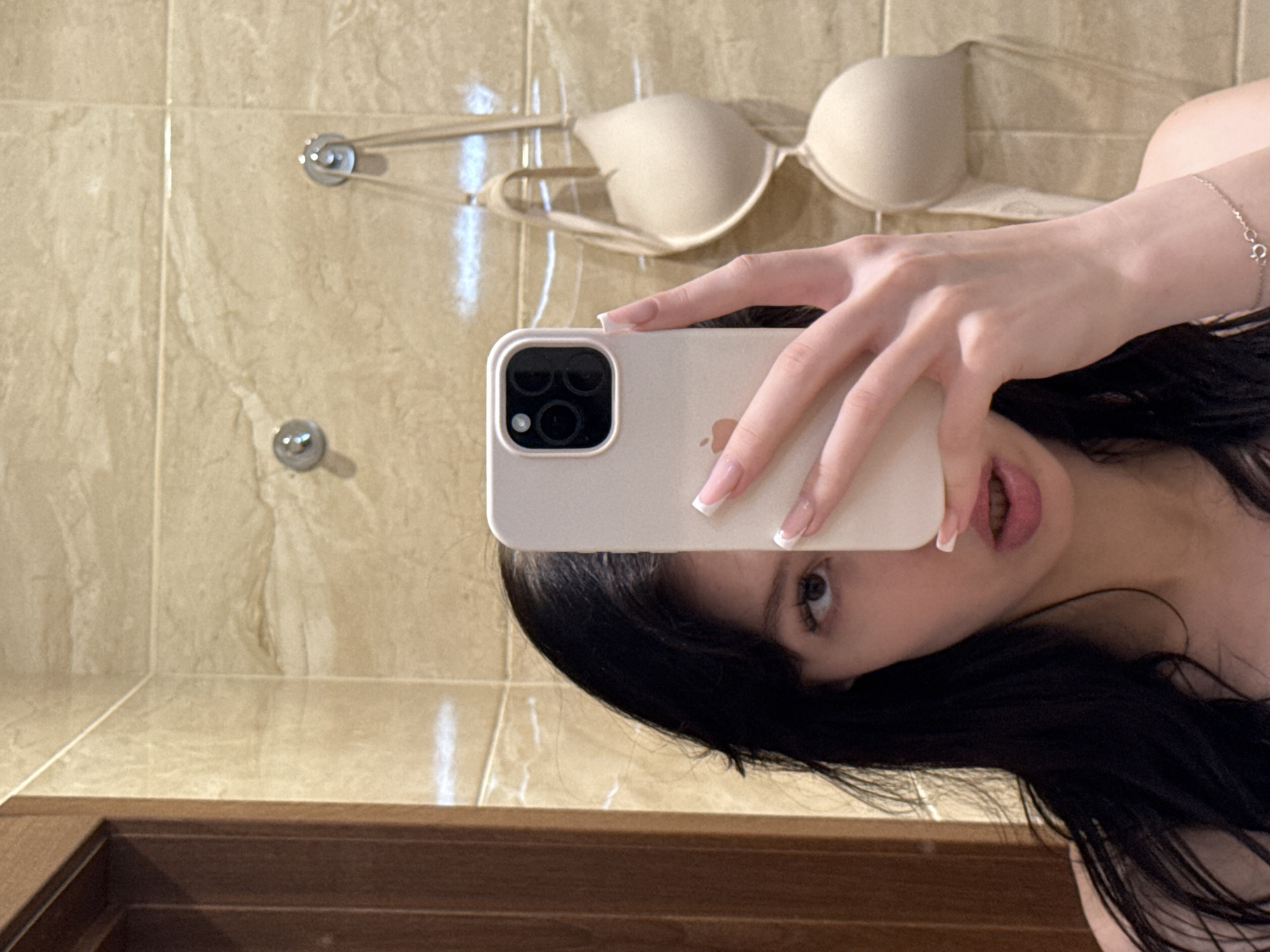 A young woman with dark hair is taking a selfie in a bathroom. She is holding an iPhone in her right hand, obscuring part of her face. A beige bra is hanging on the wall behind her, on two hooks. The bathroom walls are tiled with a beige, marble-like pattern. A wooden doorframe is visible on the left side of the image. The woman is wearing a light-colored top and a delicate silver bracelet on her left wrist.