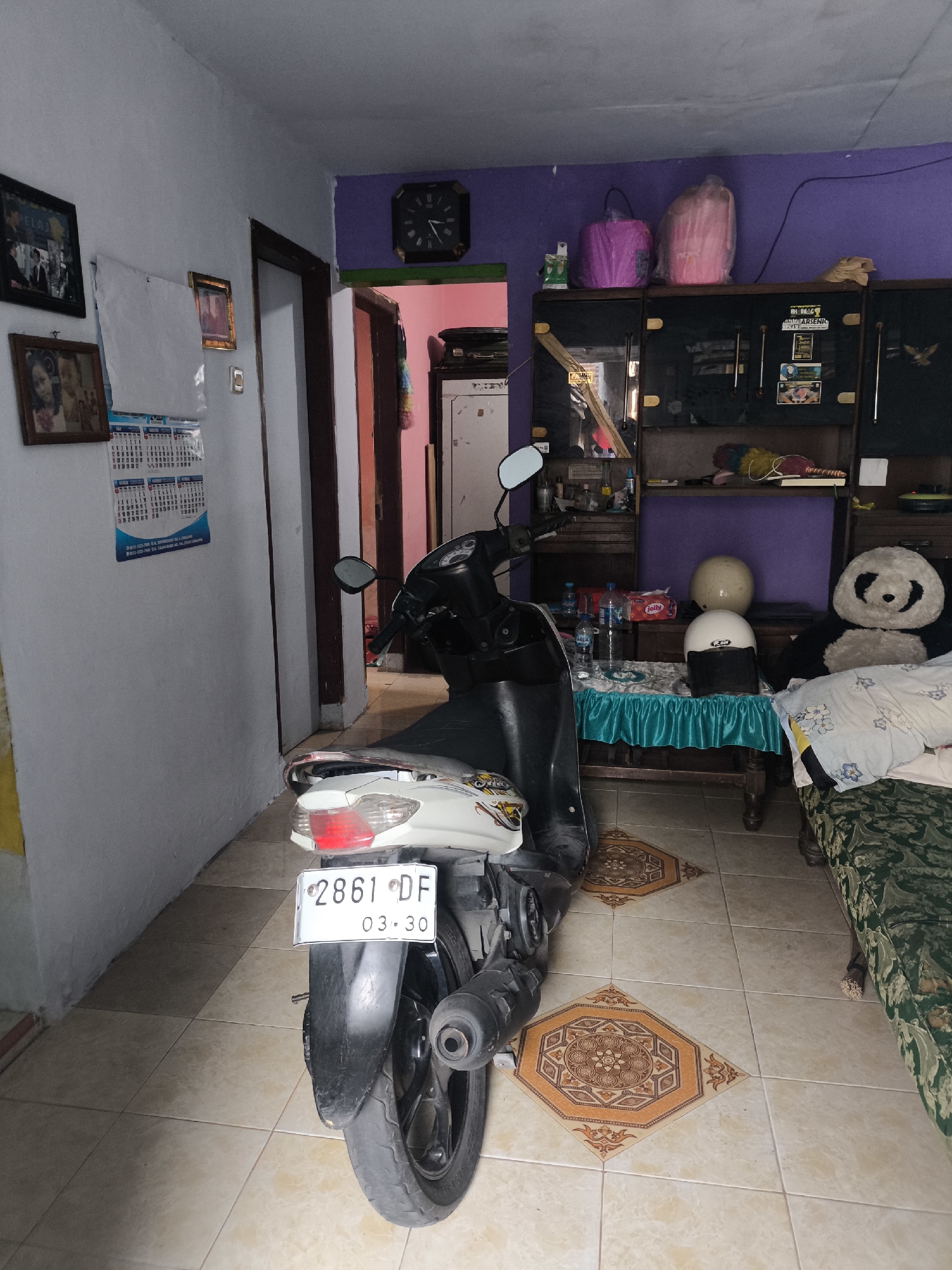 The image shows a cluttered indoor scene, possibly a living room or storage area, with a motorcycle parked centrally. The room has tiled flooring with decorative inlays. On the left, a wall is adorned with framed photographs and a calendar. To the right, a large wooden cabinet is filled with various items, including bottles, packages, and what appears to be a large stuffed panda. Two motorcycle helmets are visible on a table covered with a teal cloth, set in front of the cabinet. A partially visible sofa with a patterned green fabric is on the far right. A doorway leads to another room, where the walls are painted purple and pink, and a clock is mounted on the purple wall. The lighting is dim, with a yellowish cast. The location is identified as Citrodiwangsan, Indonesia.