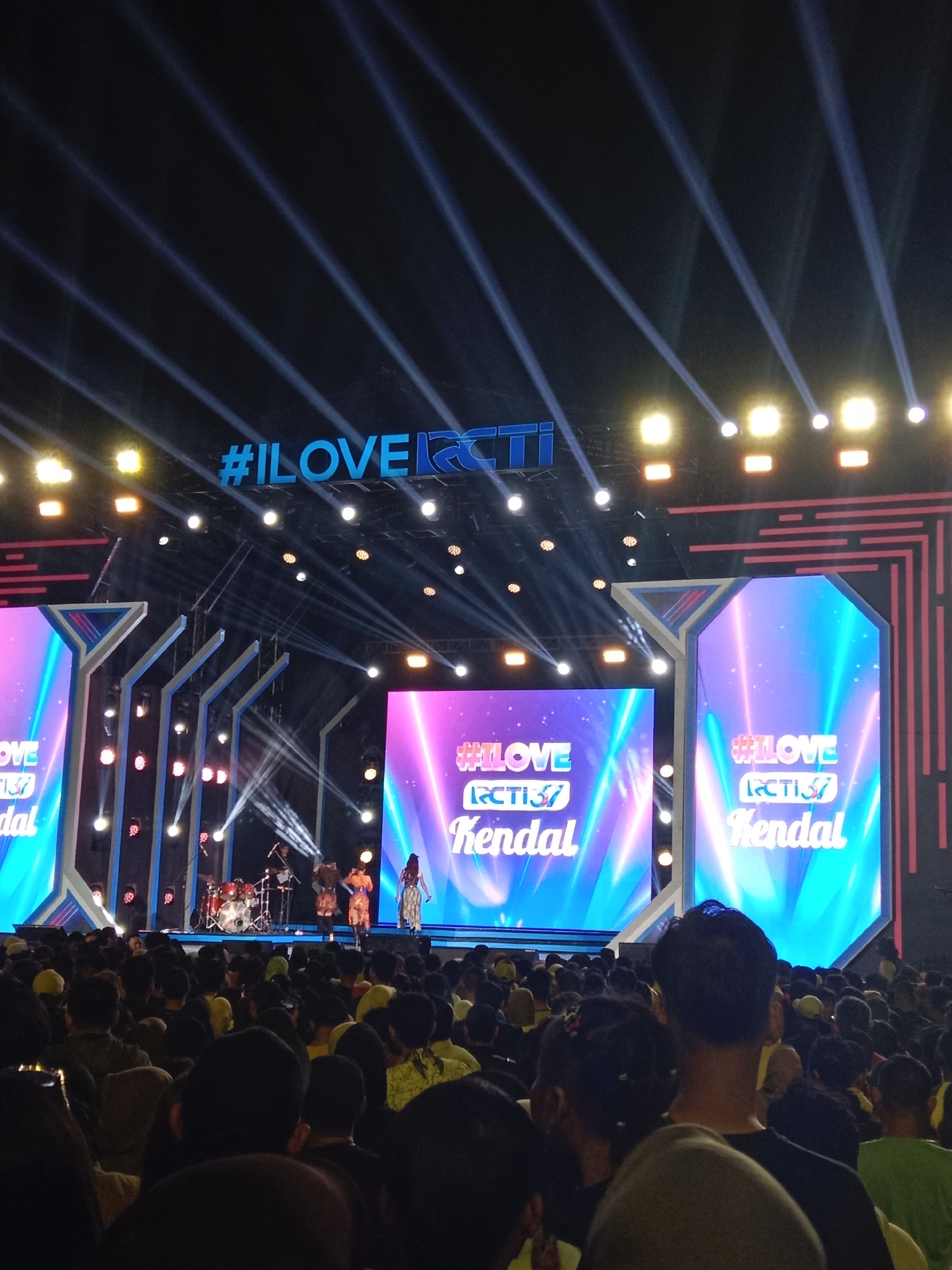 This is a photograph taken at night, outdoors, during an event in the city of Kendal, Indonesia. The scene depicts a large crowd gathered to watch a stage performance. The stage is illuminated by numerous spotlights and light beams, creating a dramatic effect against the dark sky. Two large LED screens are prominently displayed, one on the left and one on the right side of the stage, both displaying the text "#ILOVERCTI RCTI37 Kendal" in a bright, glowing font. On the stage, a band is performing, with a drummer visible at the back and singers in the foreground. The crowd in front of the stage is dense, with many people facing the stage. The overall atmosphere suggests a lively concert or festival.